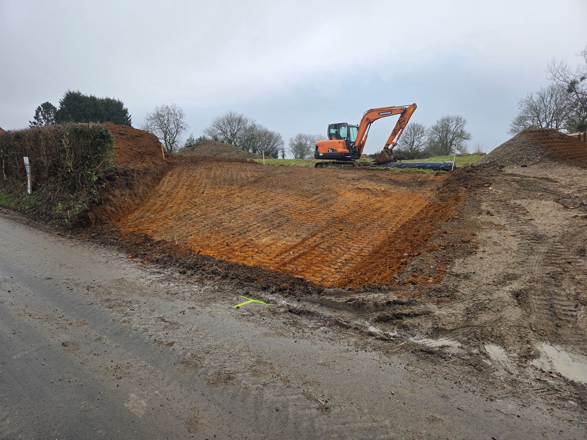 Une excavatrice sur un chantier de terrassement boueux en bord de route, creusant un grand remblai brun orangé sous un ciel couvert.