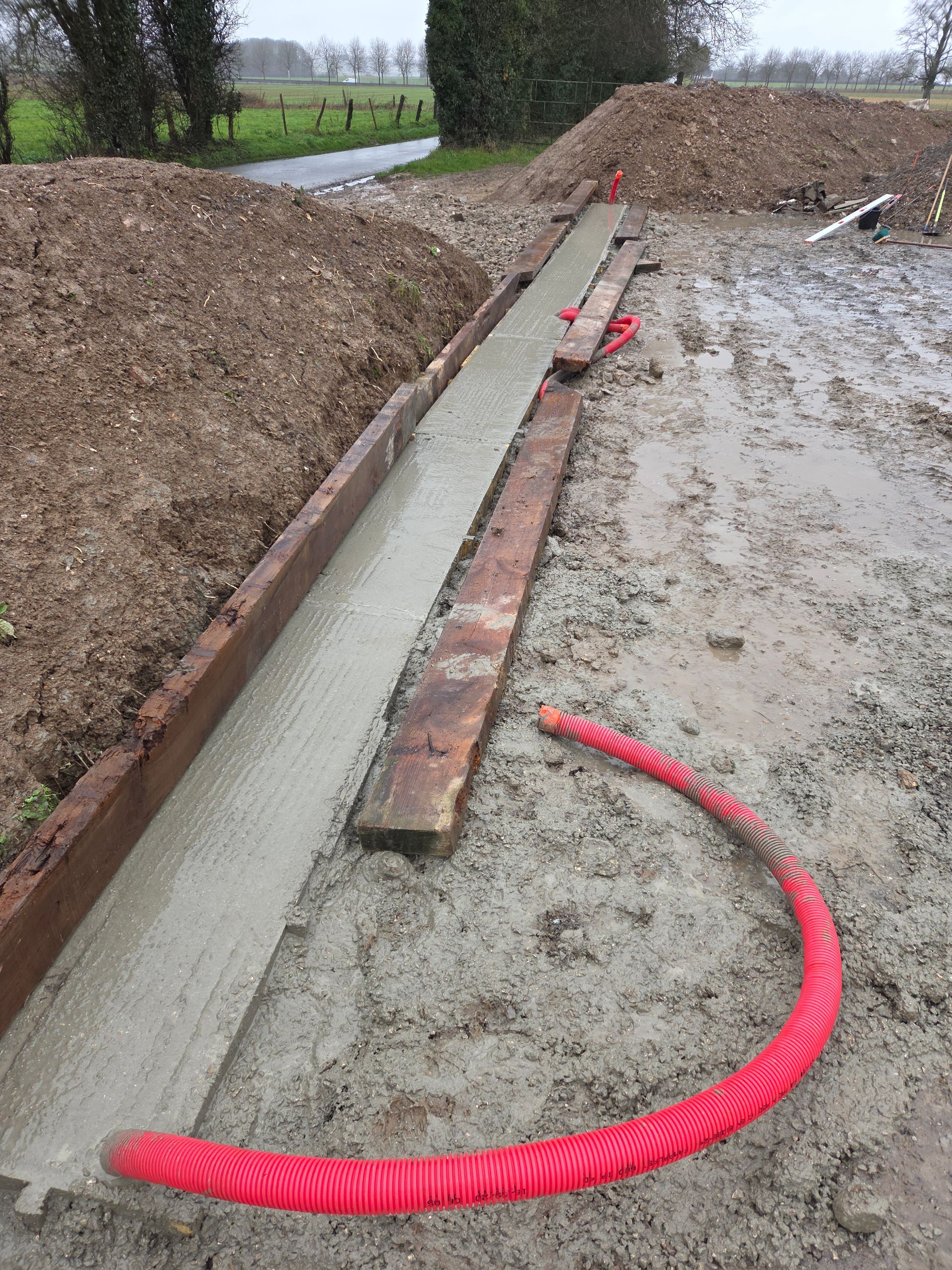 Tranchée en bord de route avec bordure en béton neuf et tuyau de drainage rouge en construction.