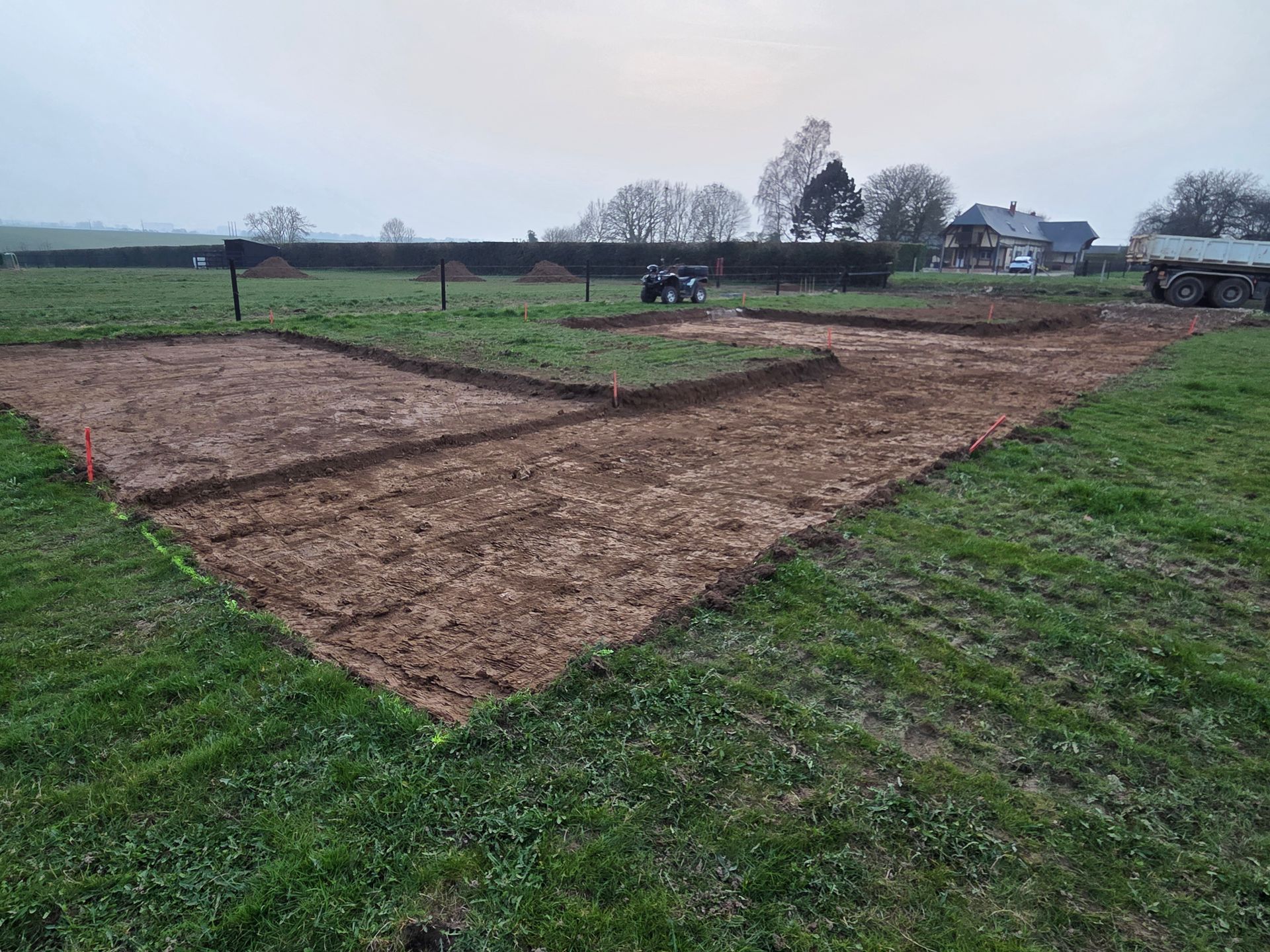 Des parcelles de jardin fraîchement labourées dans un champ herbeux sous un ciel couvert, avec des maisons au loin.