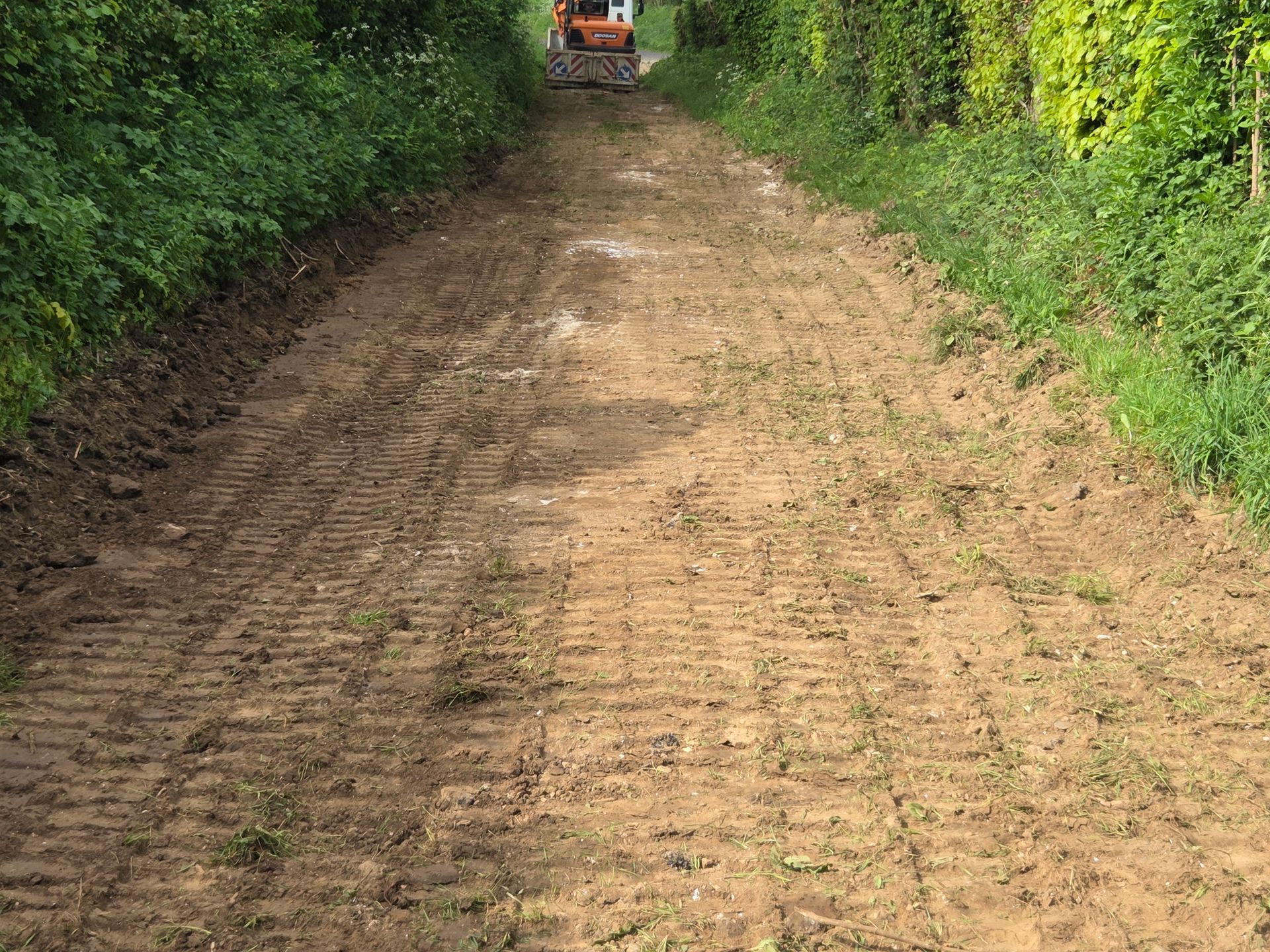 Chemin de terre traversant une dense végétation, avec un petit véhicule orange au loin.
