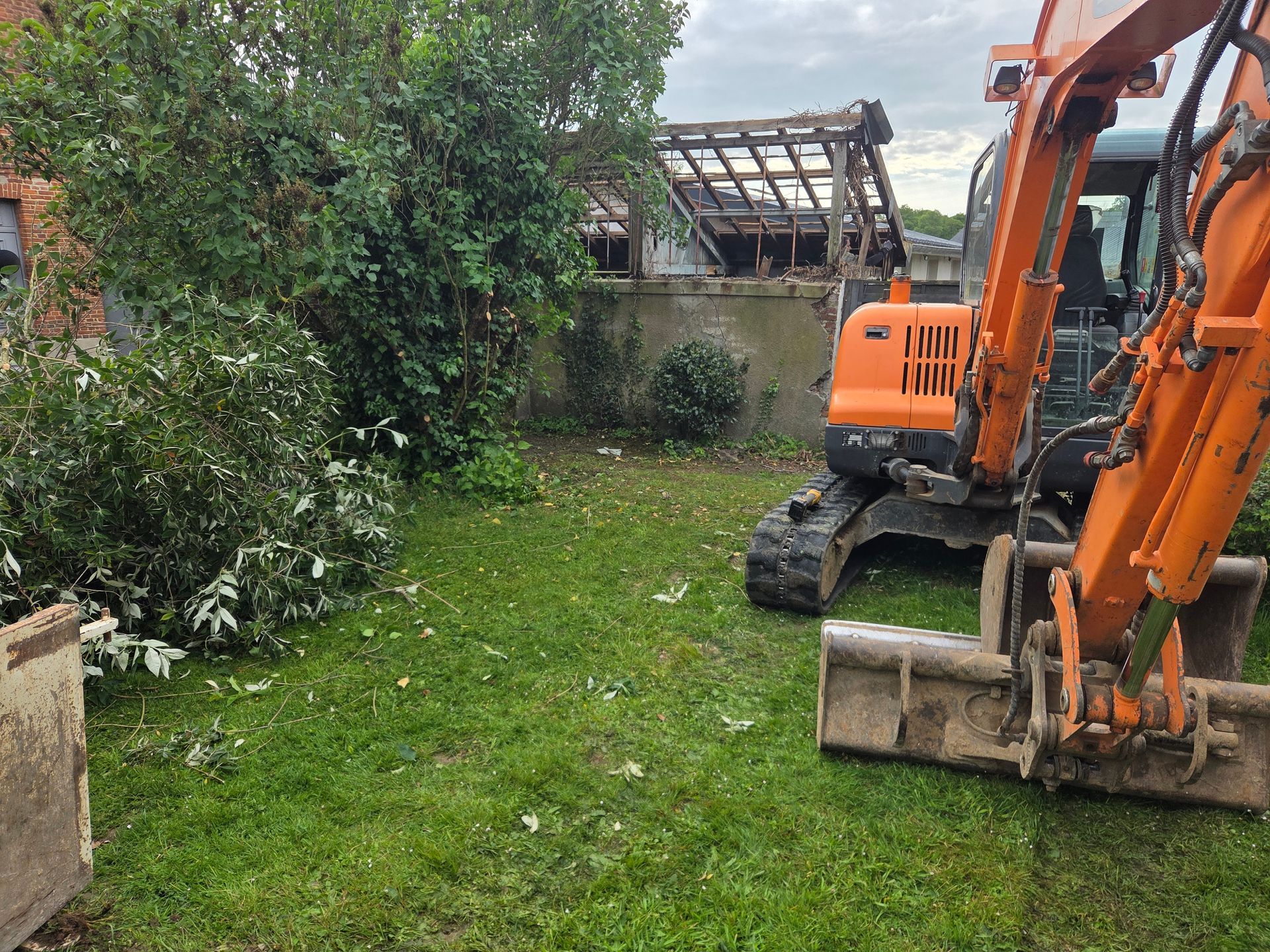 Une pelleteuse orange dans un jardin gazonné, à côté d'un arbre et d'une maison en construction.