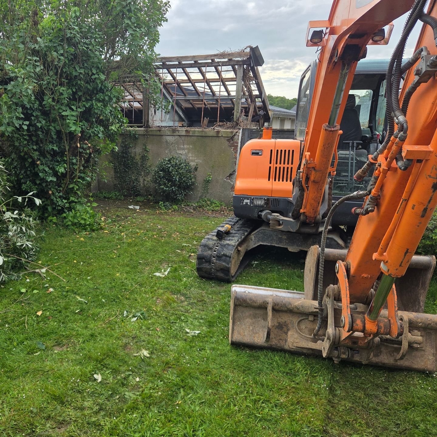 Des excavatrices orange à côté d'un bâtiment endommagé et d'une cour herbeuse pendant la démolition.