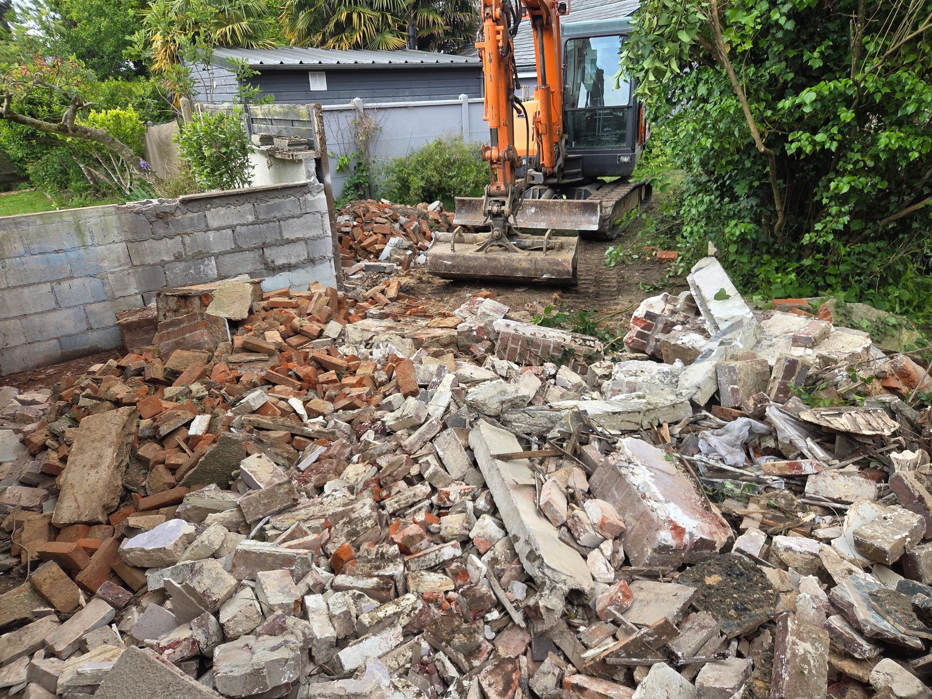 Des débris de construction entassés dans une cour, à côté d'une pelleteuse et d'un mur partiellement démoli.