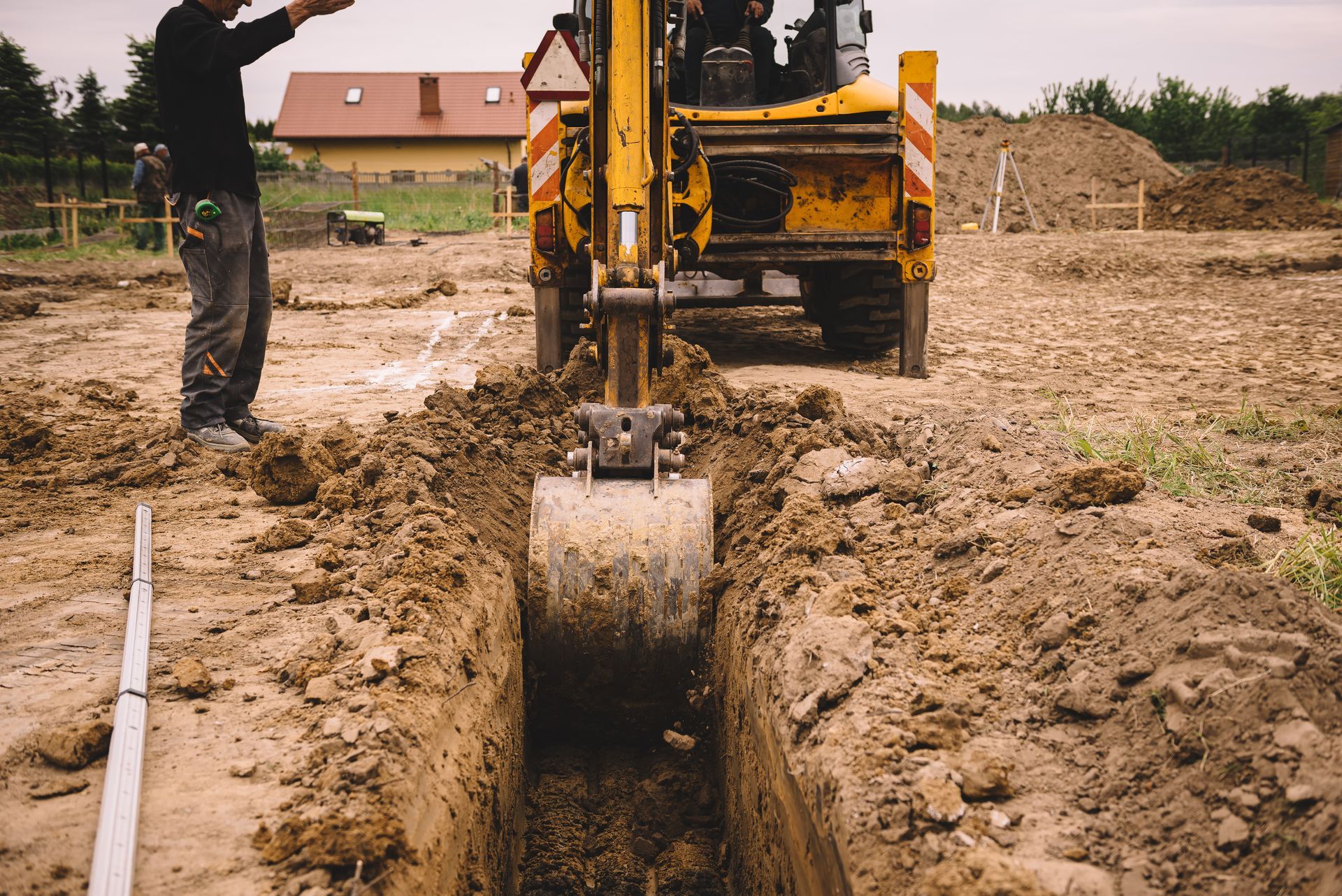 Une pelleteuse jaune creuse une tranchée sur un chantier boueux, un ouvrier se tenant à proximité.
