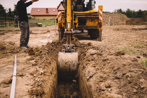 Une pelleteuse creuse une tranchée sur un chantier boueux, un ouvrier se trouvant à proximité.