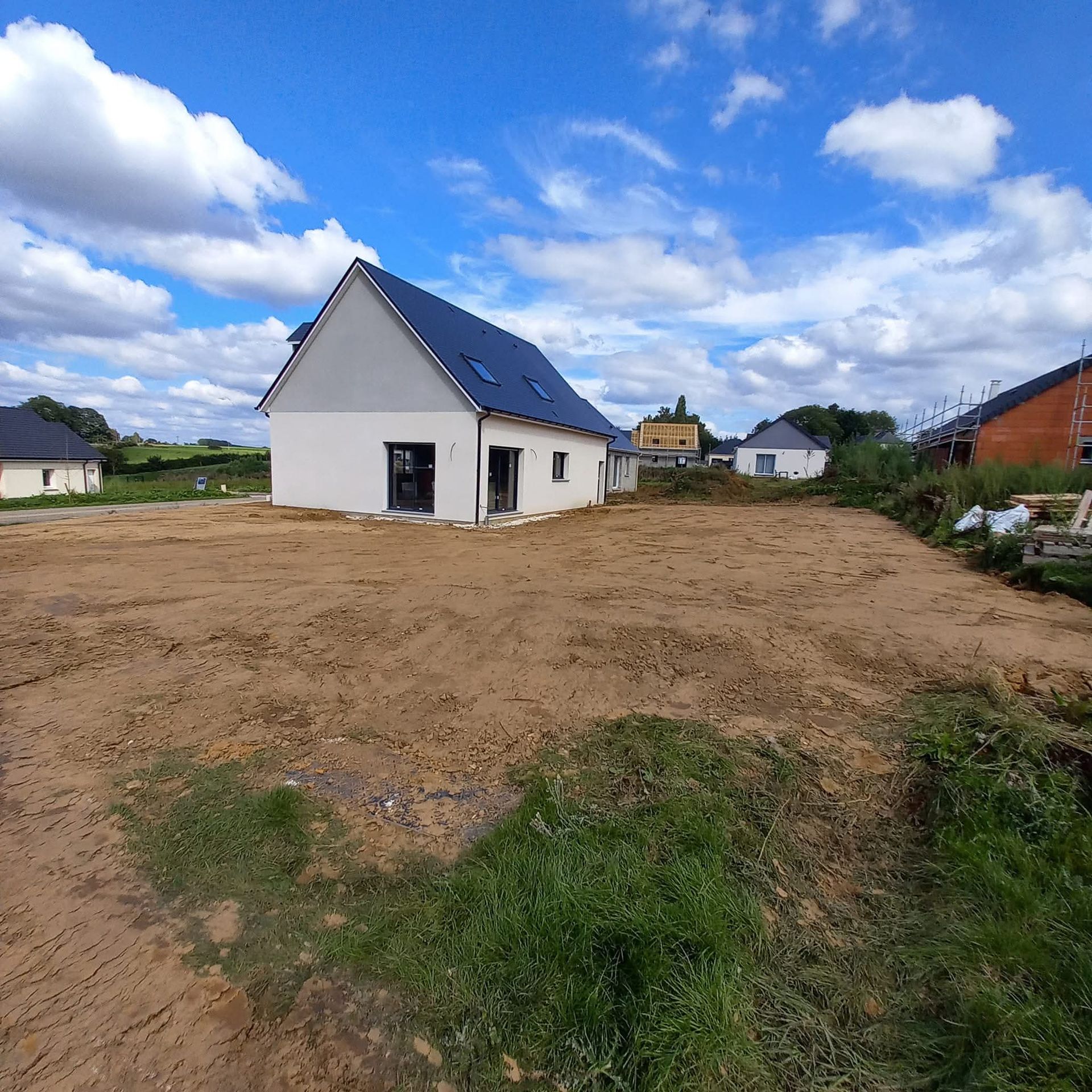Une nouvelle maison blanche est en construction sur un terrain vague sous un ciel bleu nuageux.