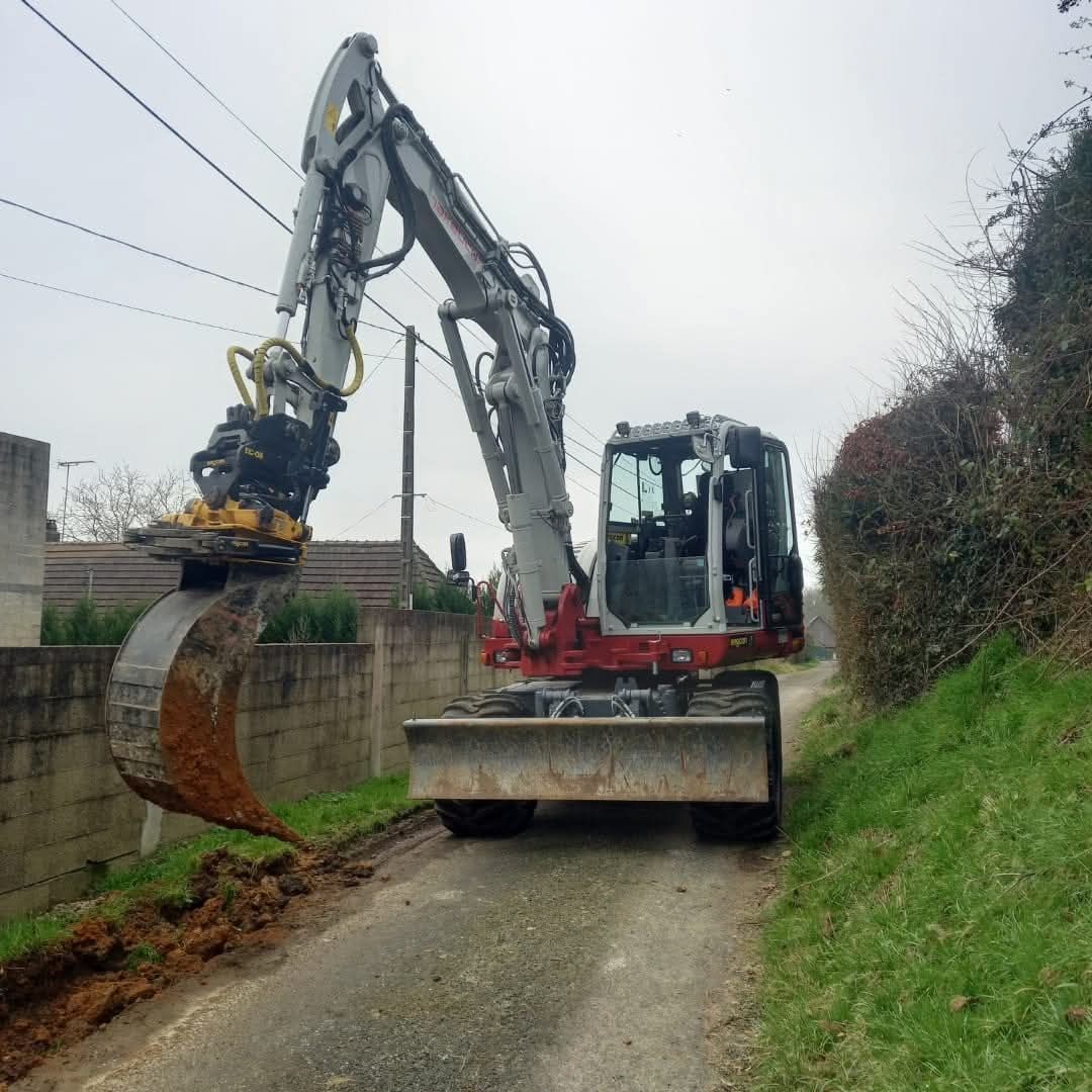 Une pelleteuse dégage la boue d'un chemin de campagne étroit, longeant un mur et une haie.