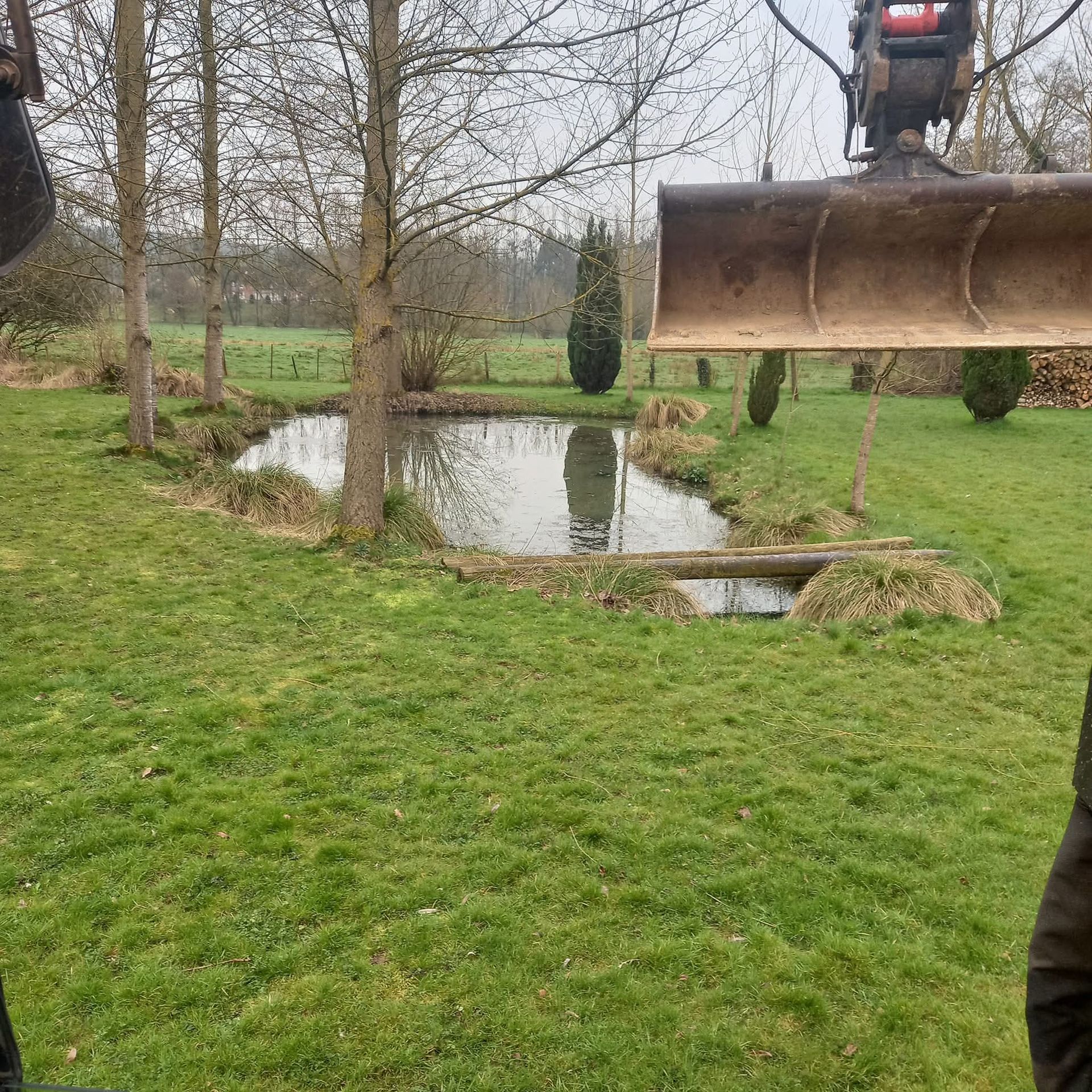 Une cour herbeuse avec un petit étang et des arbres dénudés, vue à côté d'un godet de chantier sur la droite.
