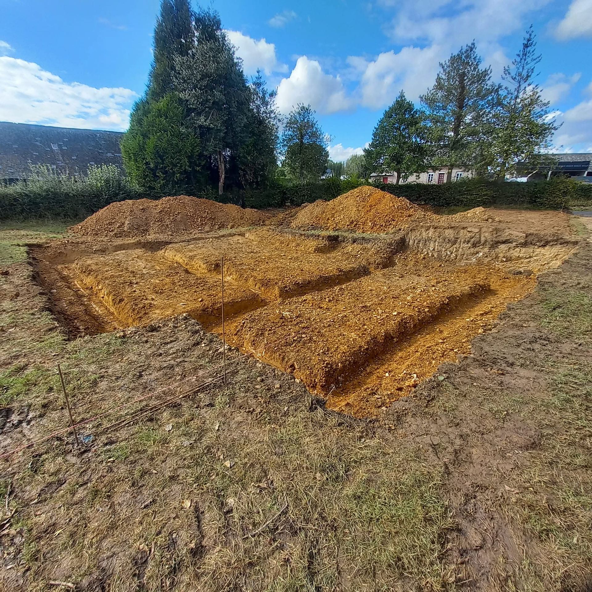 Des plates-bandes rectangulaires fraîchement creusées dans un champ de terre, avec des monticules de terre, des arbres et un ciel bleu en arrière-plan.