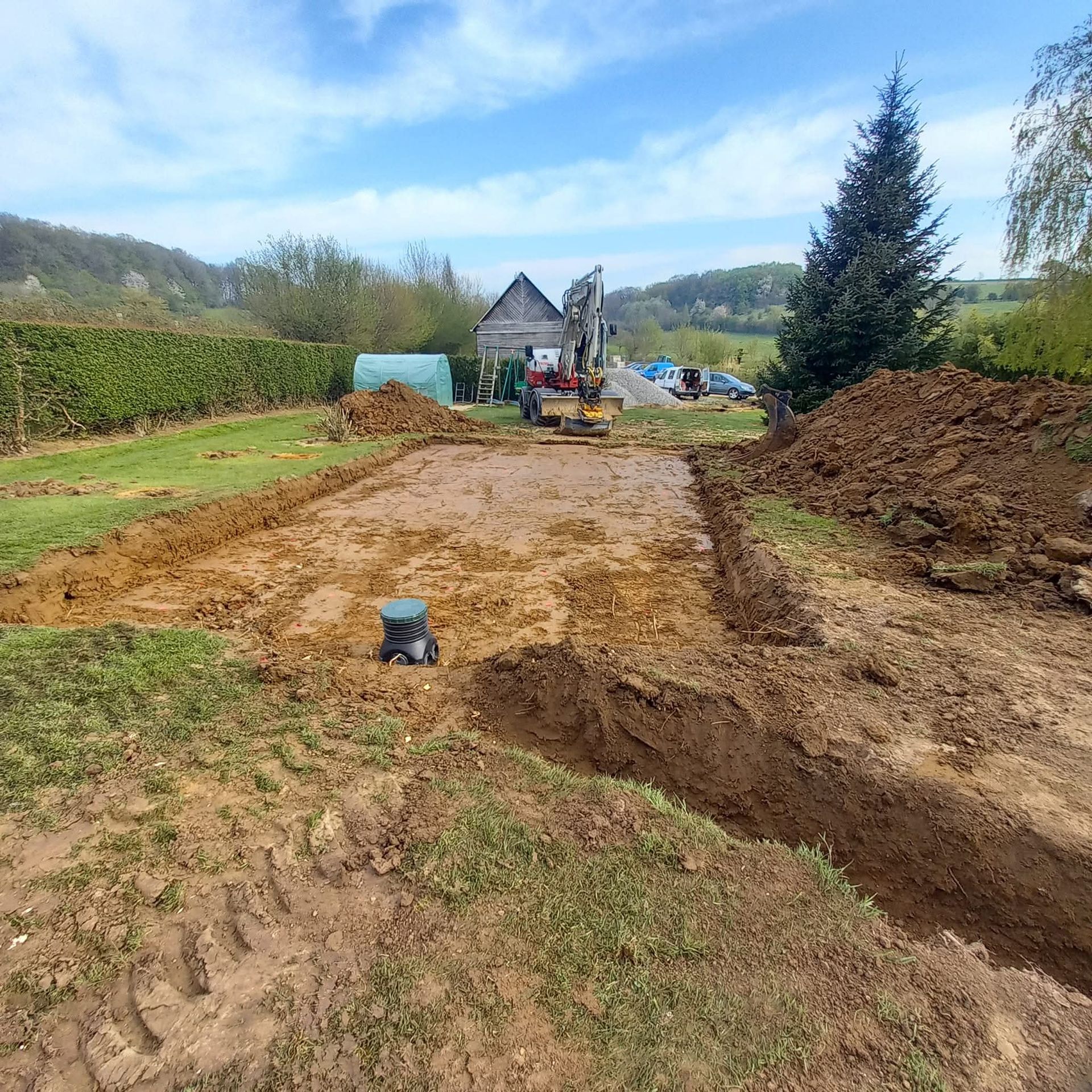 Un ouvrier en pantalon bleu utilise un tuyau d'arrosage pour nettoyer un trou de drainage herbeux.