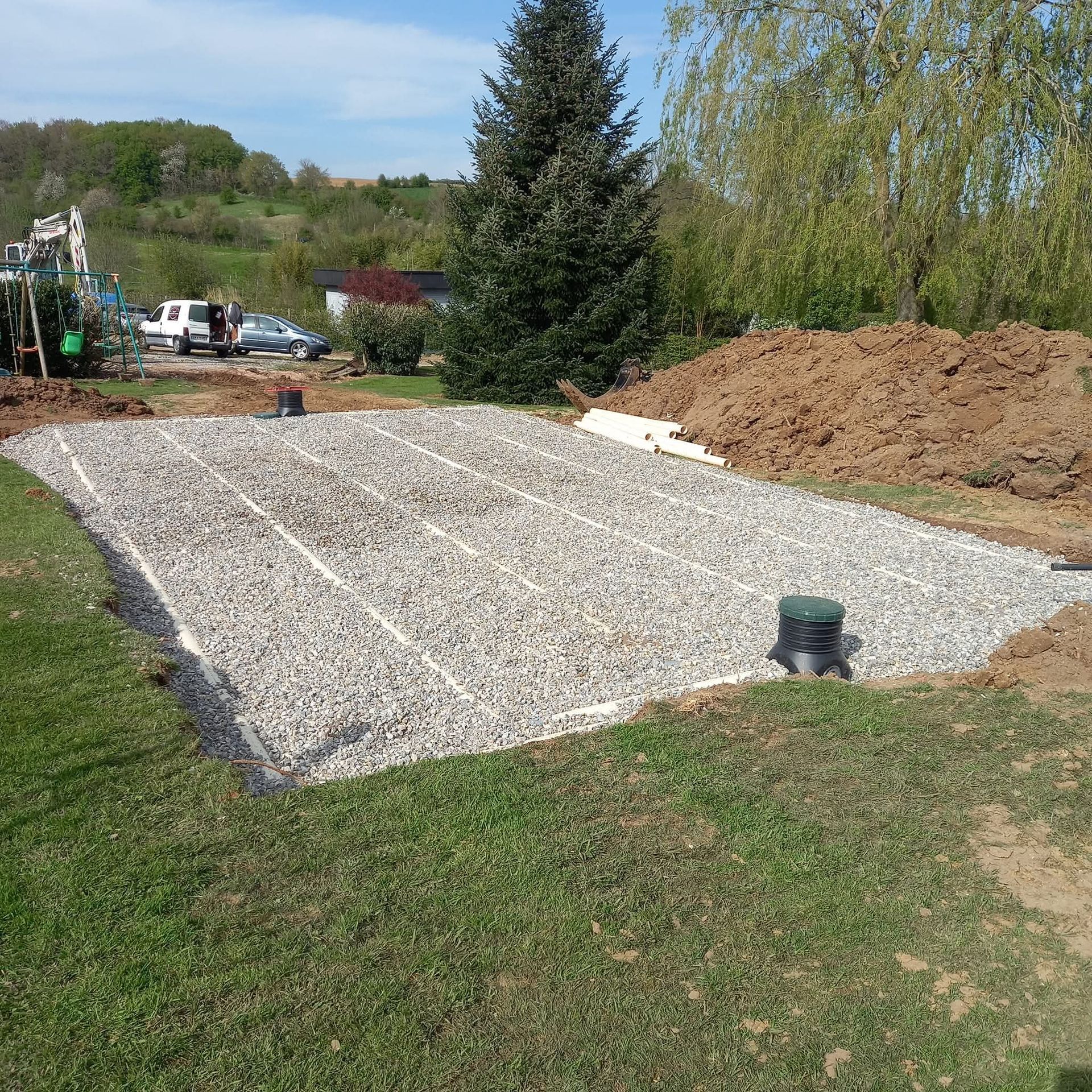 Terrain en pente recouvert de gravier, bordé de pelouse, avec des tas de terre, des arbres et un petit tuyau au premier plan.