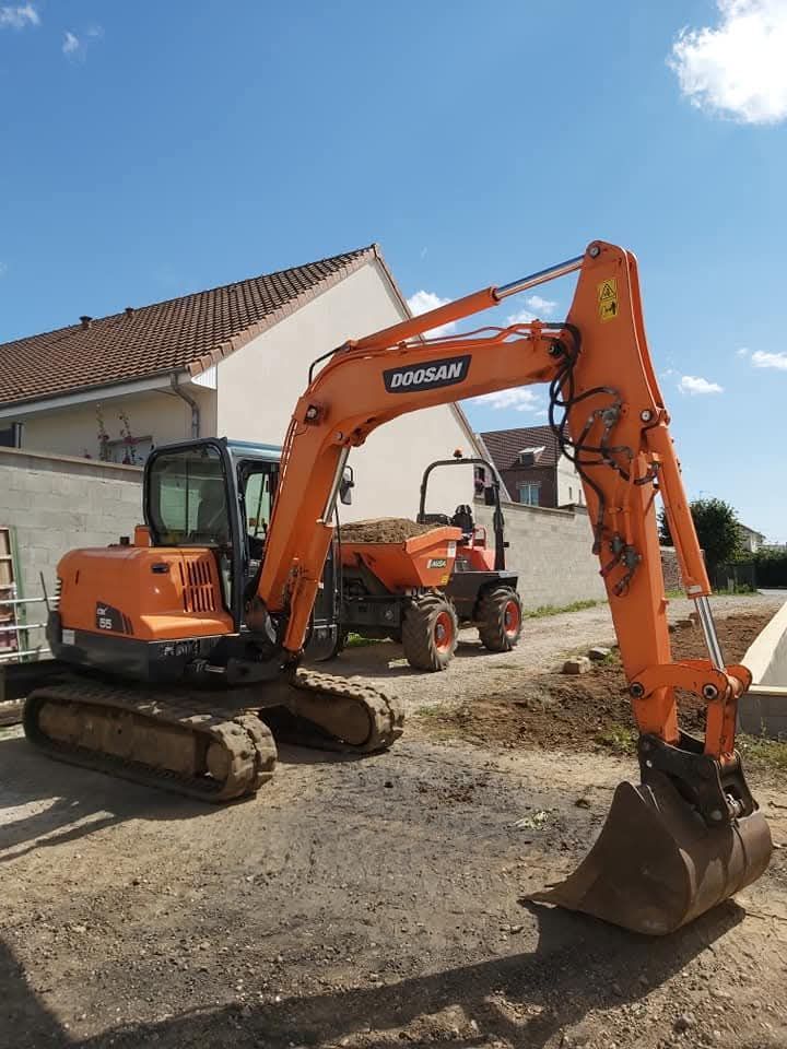 Une pelleteuse orange est garée sur du gravier à côté d'une maison, avec une autre machine en arrière-plan.