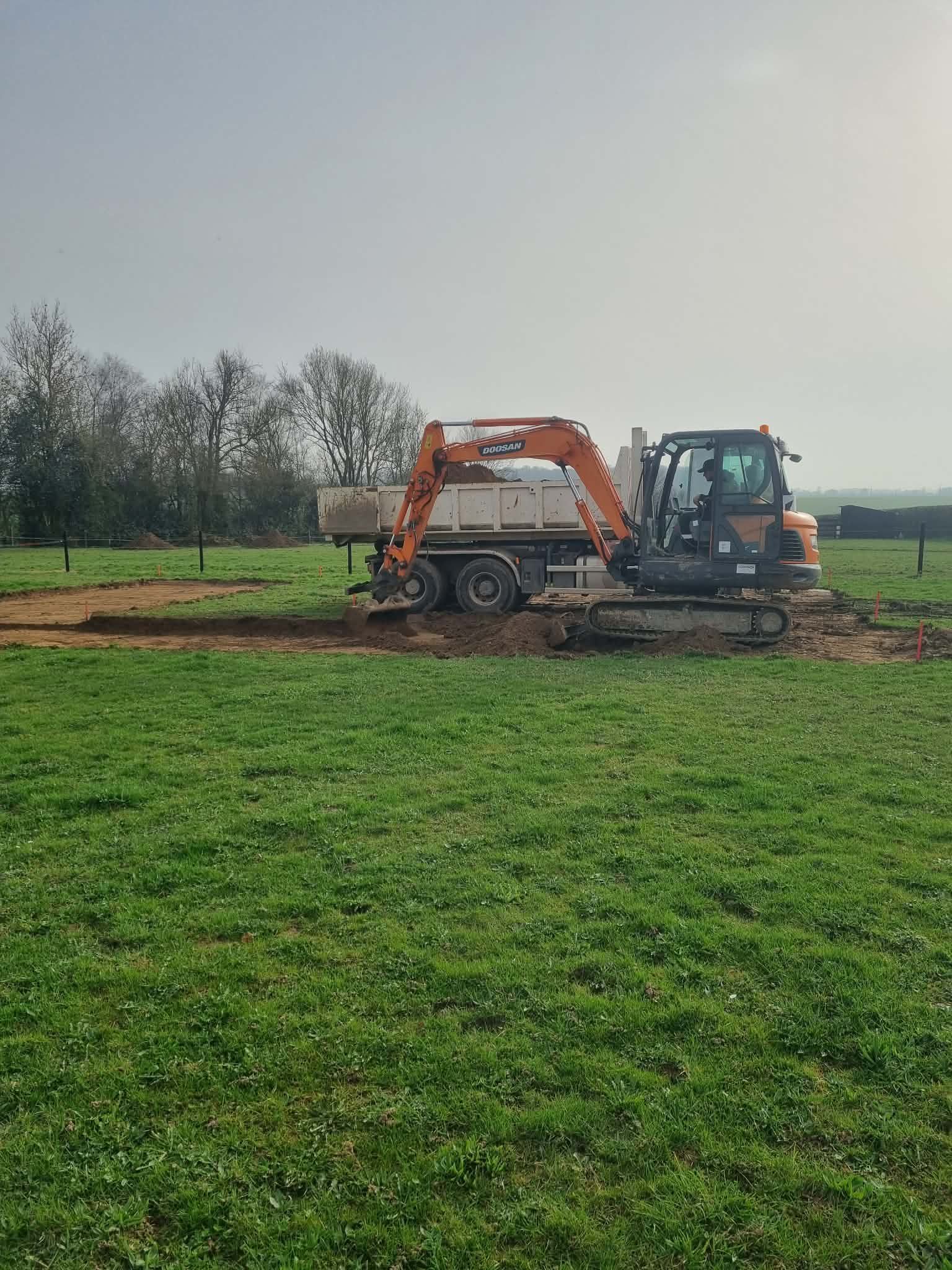 Une pelleteuse orange charge de la terre sur un camion dans un champ boueux sous un ciel couvert.