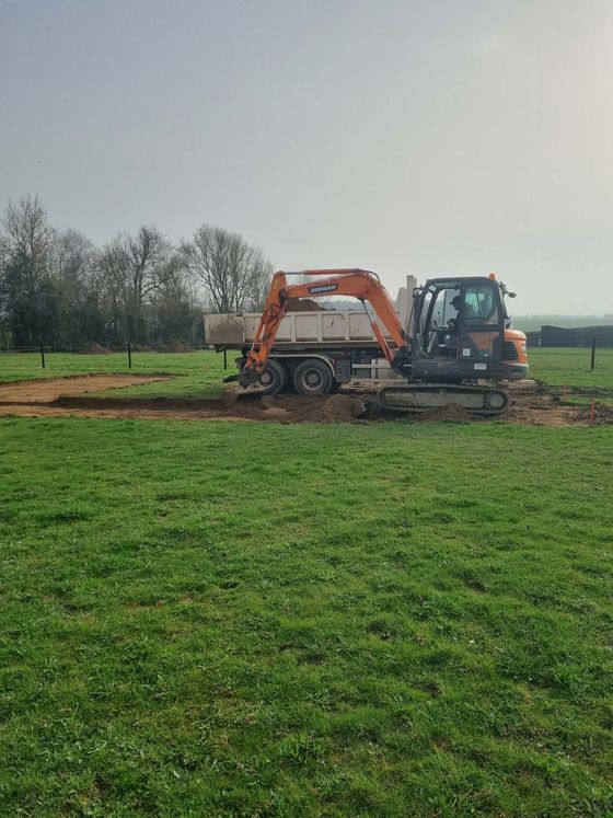 Une pelleteuse orange et un tracteur bleu travaillent sur un terrain boueux dans un champ herbeux sous un ciel gris.
