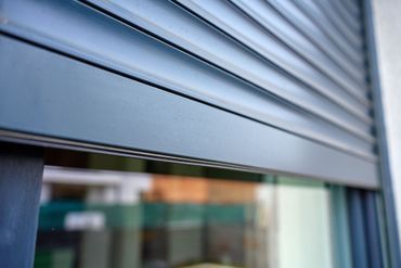 Gros plan sur un volet roulant métallique gris avec une vitrine floue derrière.