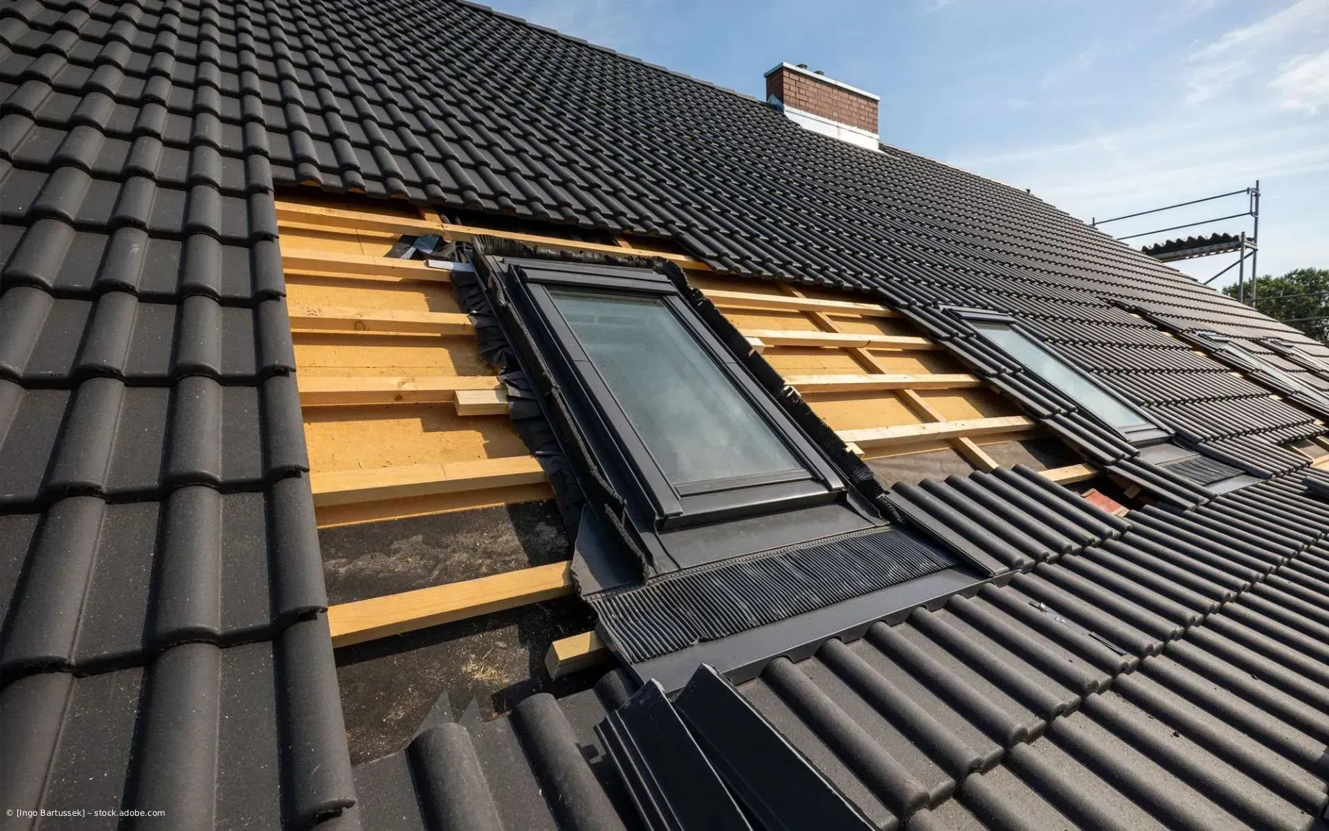 Dachfenster im Einbau auf einem dunklen Ziegeldach, umgeben von freiliegenden Holzlatten.