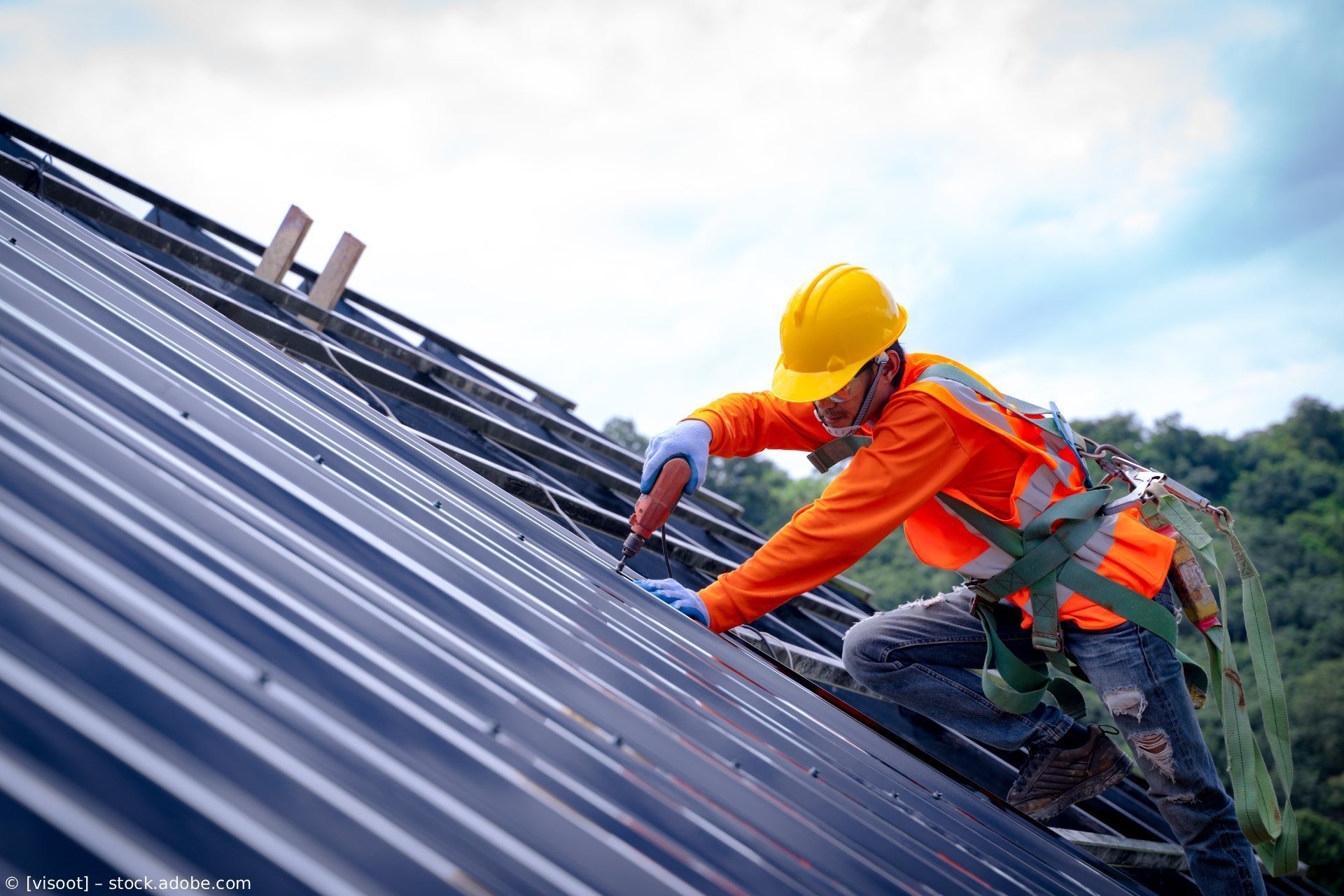 Arbeiter in orangefarbener Schutzkleidung und gelbem Schutzhelm sichert Metalldachplatten auf einem steilen Dach