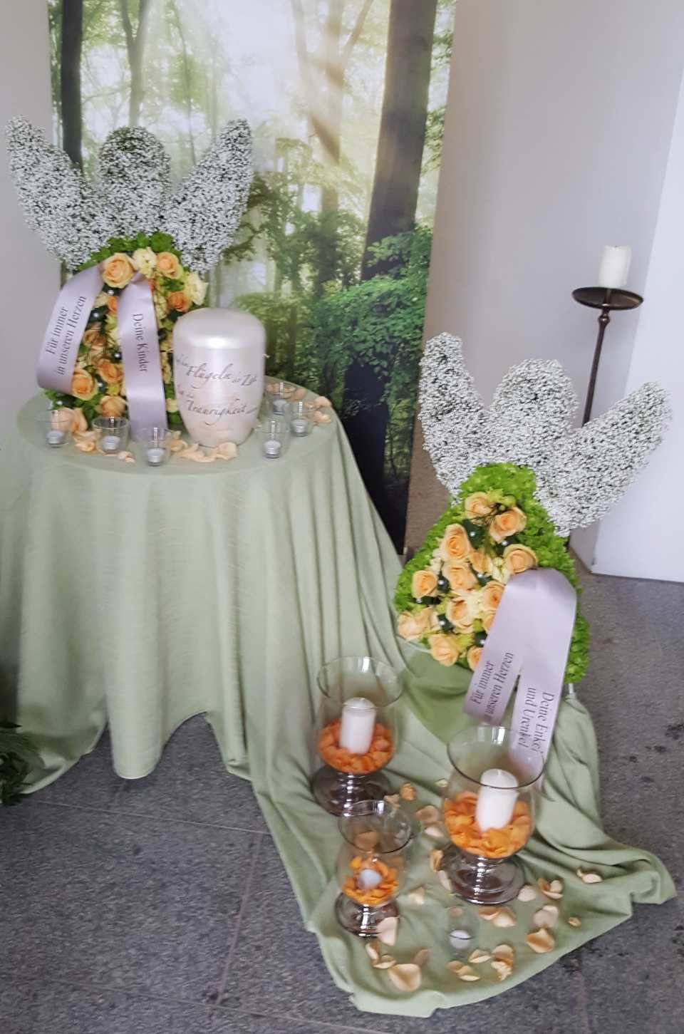 Trauergestecke mit Blumen, Urne, Kerzen und engelsförmigen Blumenarrangements; ruhige Grün- und Gelbtöne.