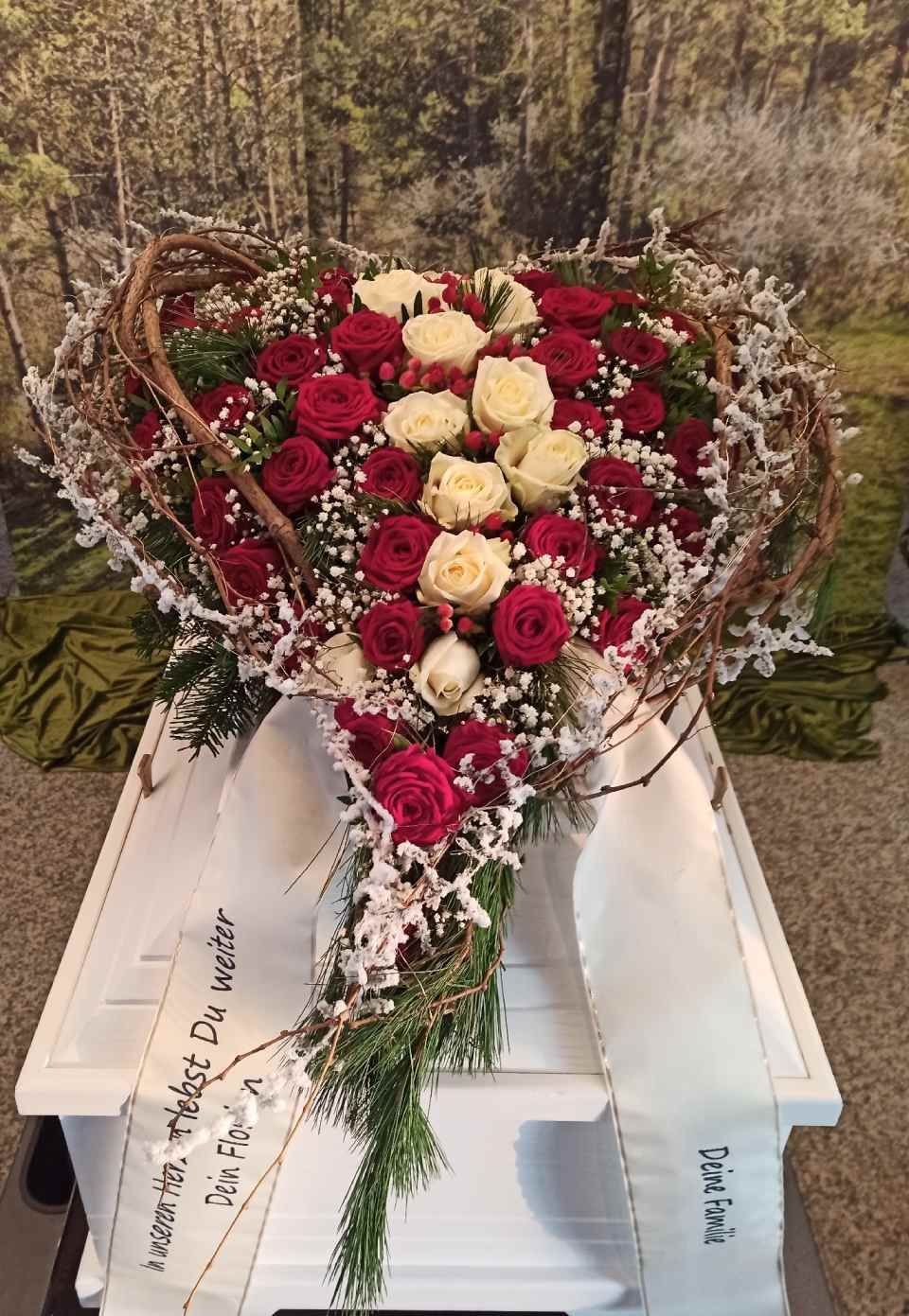 Herzförmiges Blumenarrangement aus roten und weißen Rosen auf einem weißen Sarg, mit Schleife und Grünzeug.