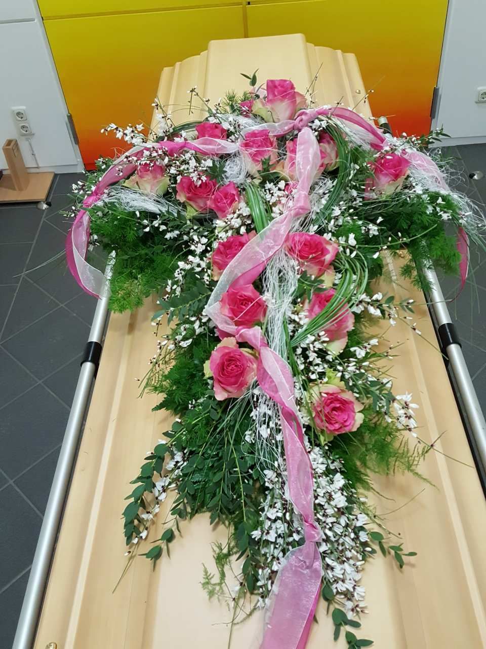 Blumenarrangement aus rosa Rosen und Grünzeug auf einem hellen Sarg.