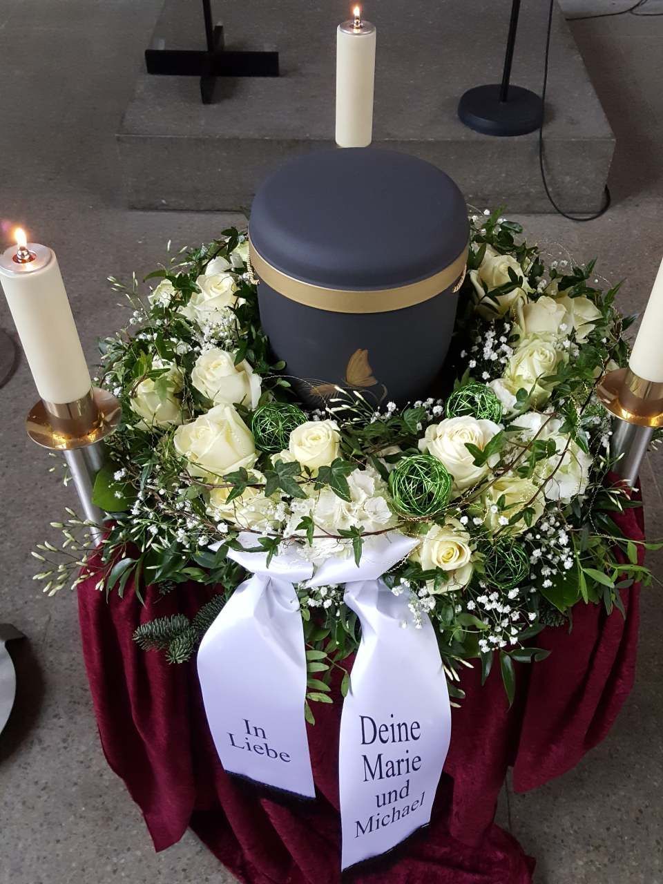Urn with floral wreath, candles, and ribbon reading 