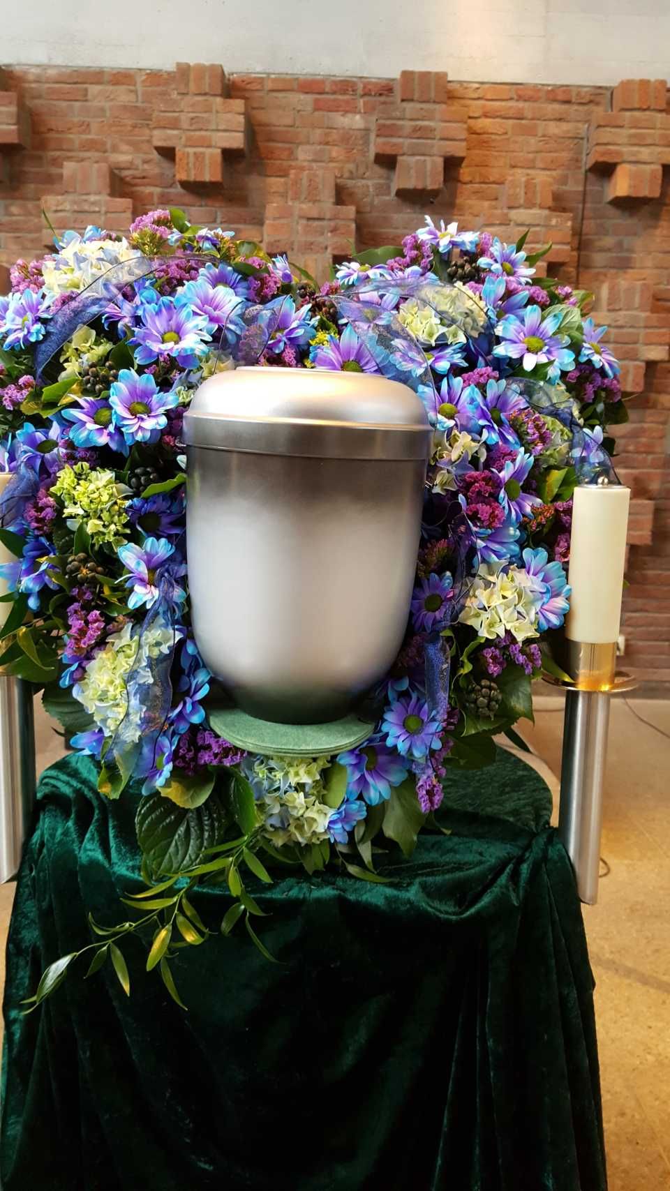 Eine Urne, umgeben von einem floralen Herzarrangement, steht auf einem drapierten grünen Tuch vor einer Backsteinmauer.