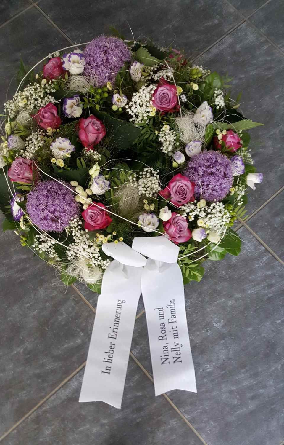 Kranz aus lila und rosa Blüten mit weißem Band; Inschrift sichtbar.