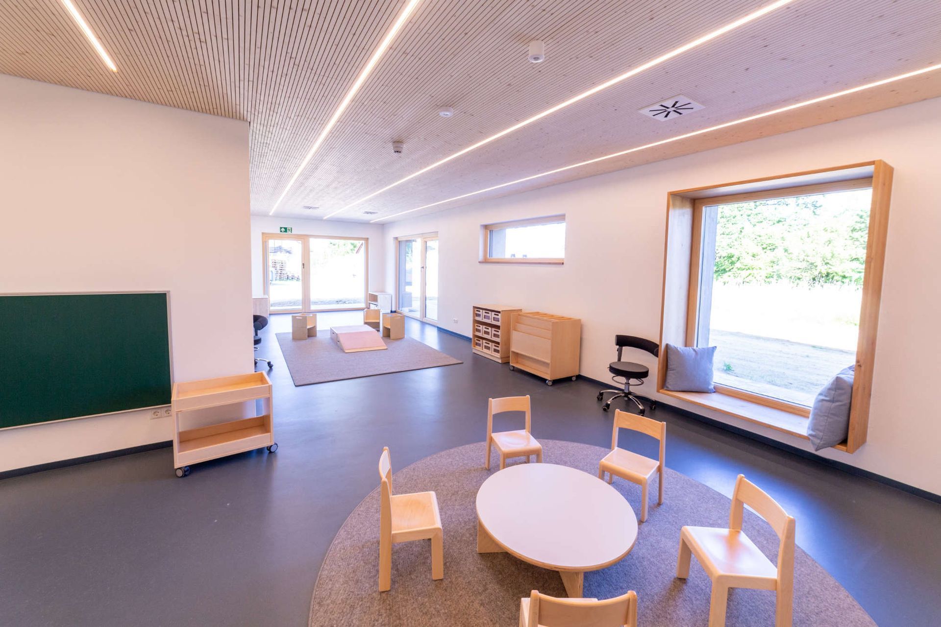 Raumgestaltung mit Kinderstühlen rund um einen runden kleinen Tisch und kleine Schränke und große Fenster mit Sitzkissen