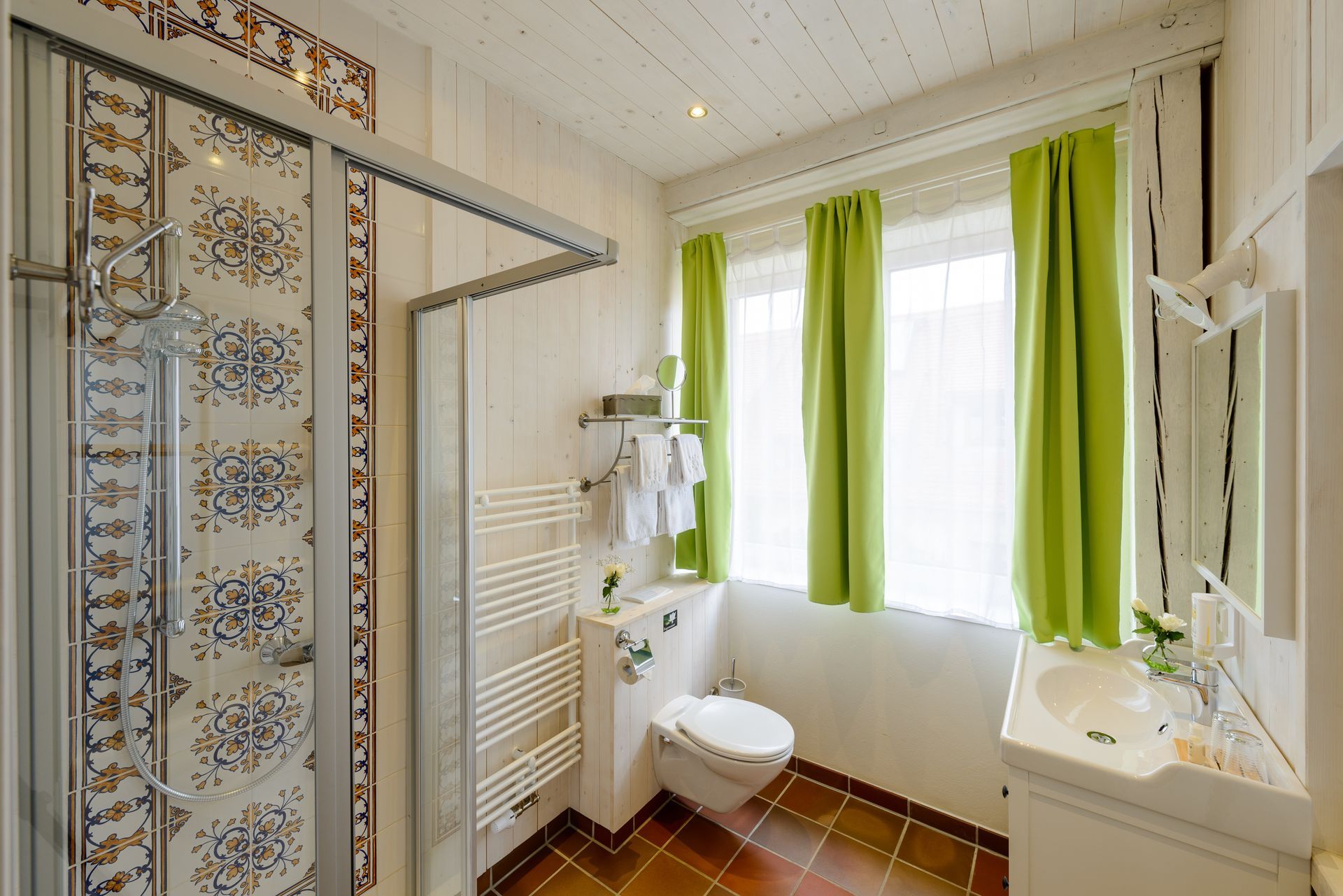 Badezimmer mit gemusterter Duschwand, weißen Armaturen, grünen Vorhängen und gefliestem Boden.