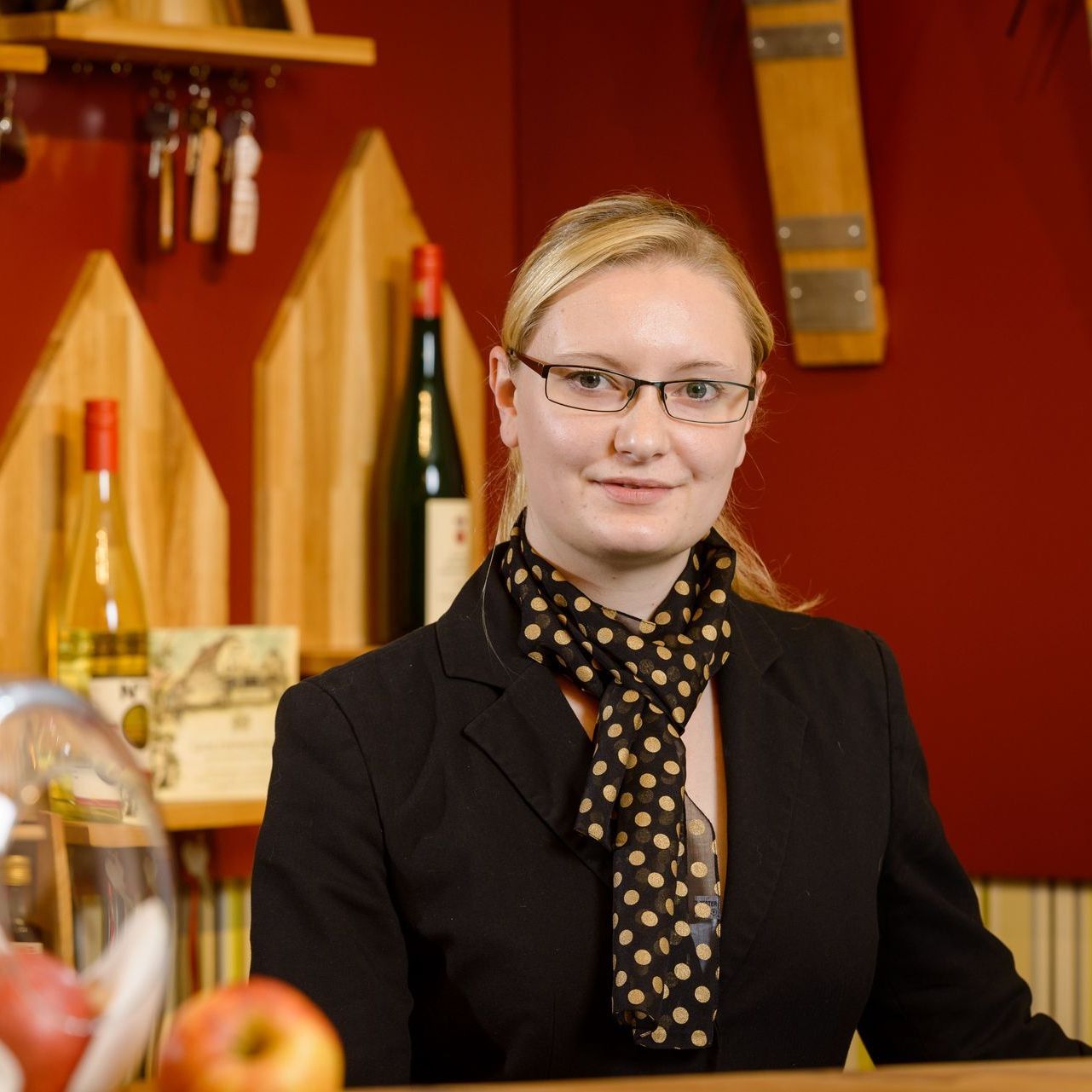 Eine Frau mit Brille und schwarzem Blazer steht vor einer Bar mit Flaschen und trägt einen gepunkteten Schal.