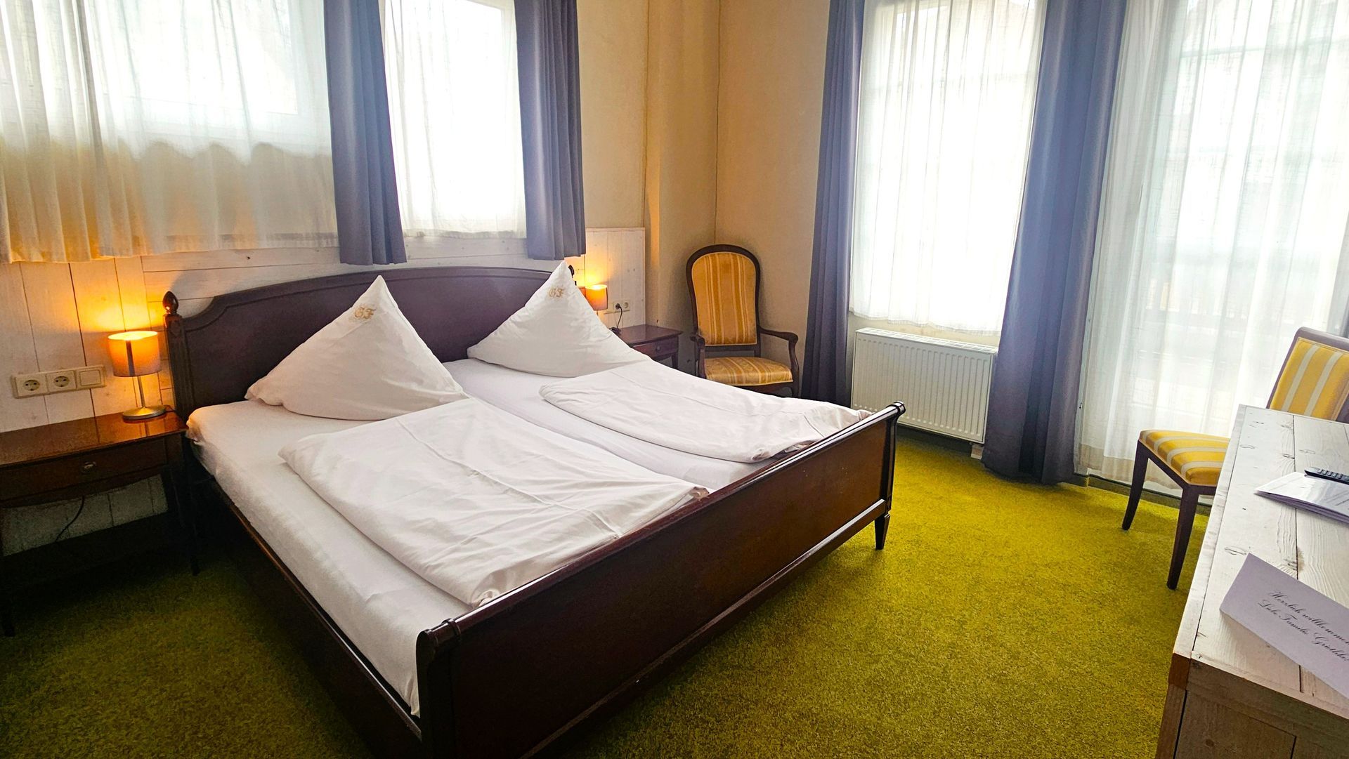 Schlafzimmer mit Doppelbett, zwei Kissen, zwei Fenstern, goldenem Stuhl und kleinem Tisch. Grüner Teppich, hellbraune Wände.