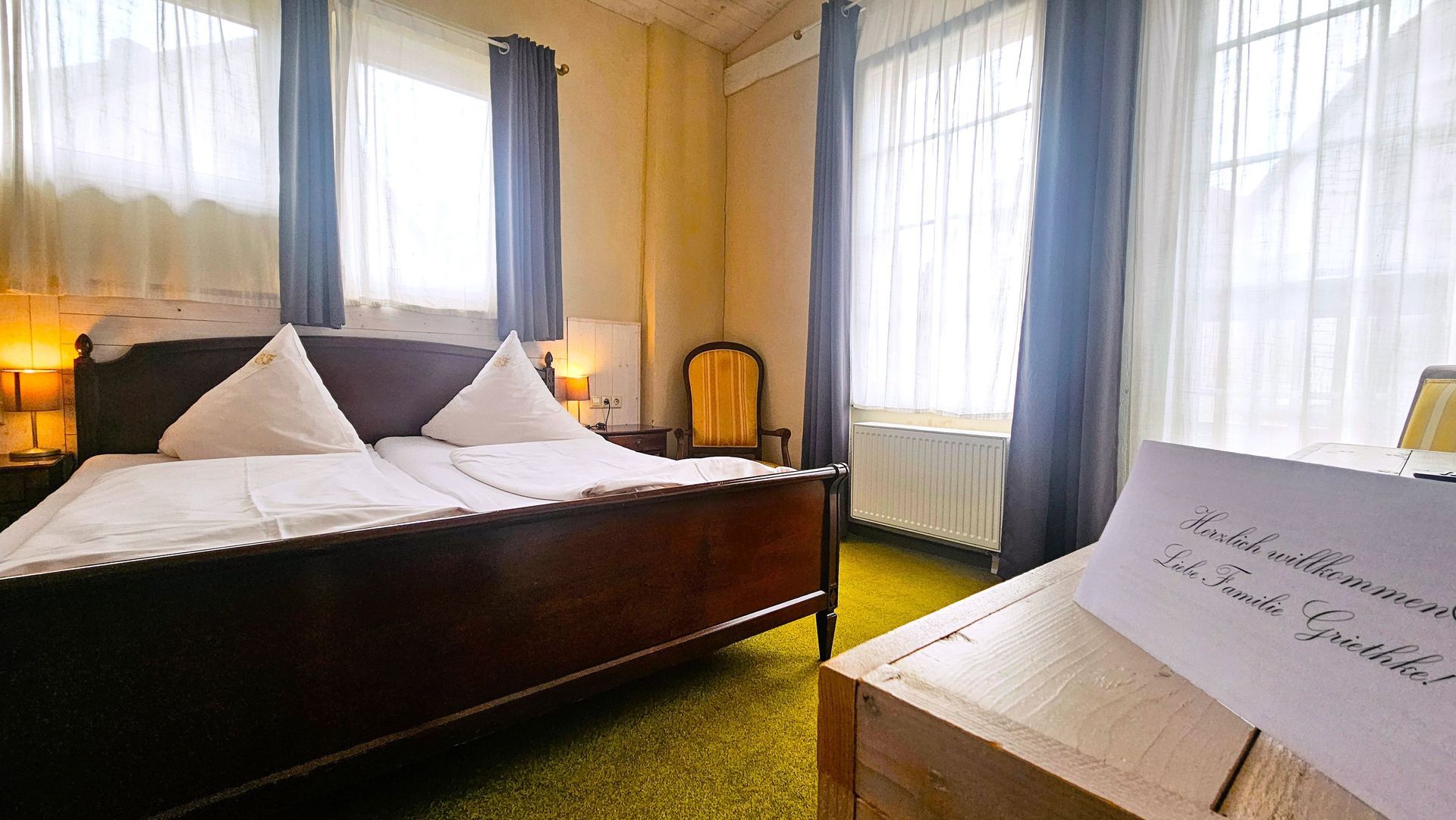 Schlafzimmer mit einem Holzbett, weißen Kissen, durchsichtigen Vorhängen und einem Schreibtisch mit einer handgeschriebenen Notiz.