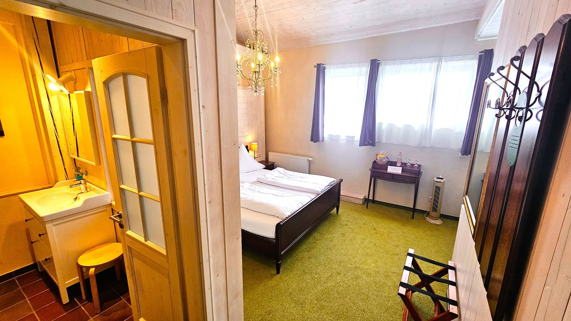 Schlafzimmerinterieur mit Bett, Badezimmertür und Garderobe. Grüner Teppich, weiße Wände und ein Kronleuchter.