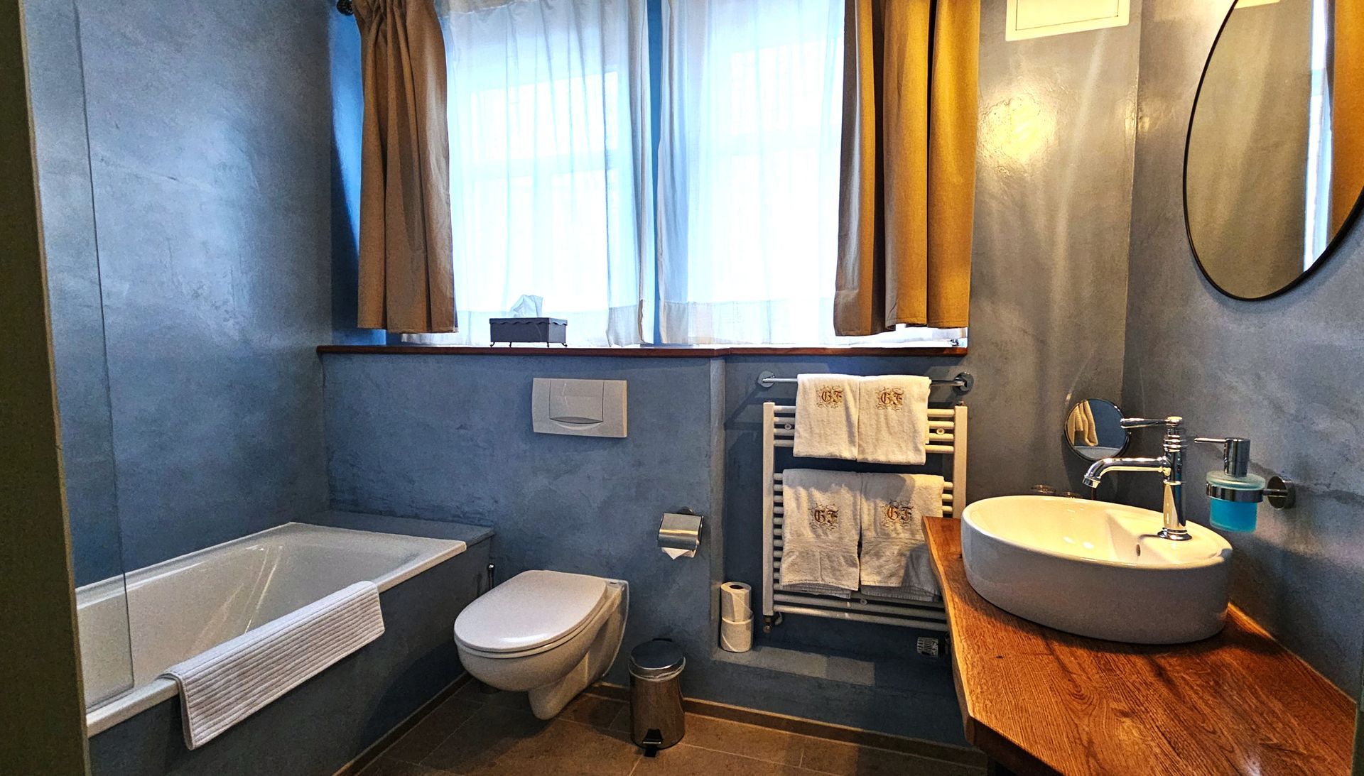 Badezimmer mit blauen Wänden, Badewanne, Toilette, Waschbecken und Fenster mit Vorhängen.