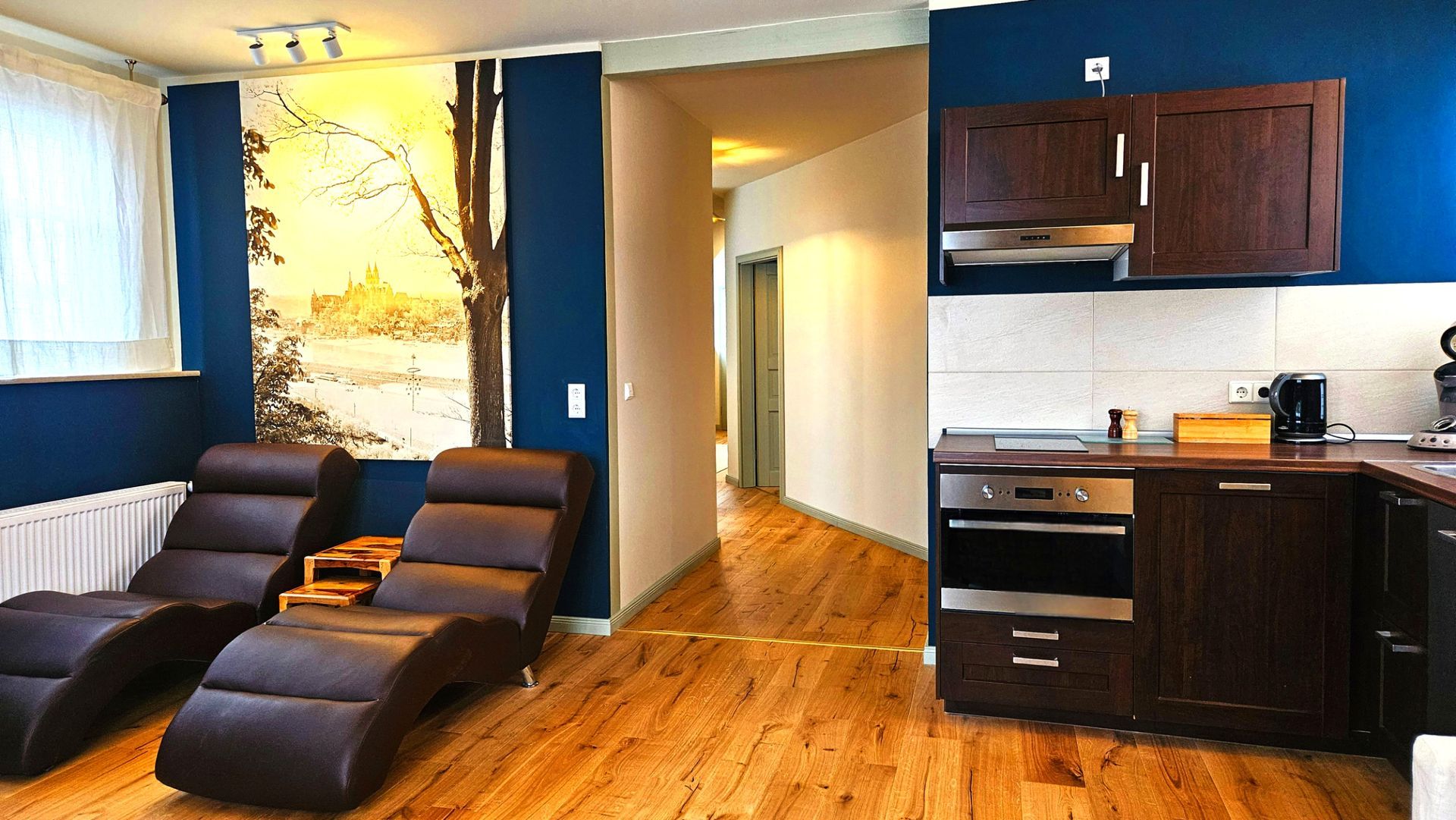 Wohnbereich mit Loungesesseln, Küche und Kunstwerken an der Wand. Dunkelblaue Wände, Holzböden und -schränke.