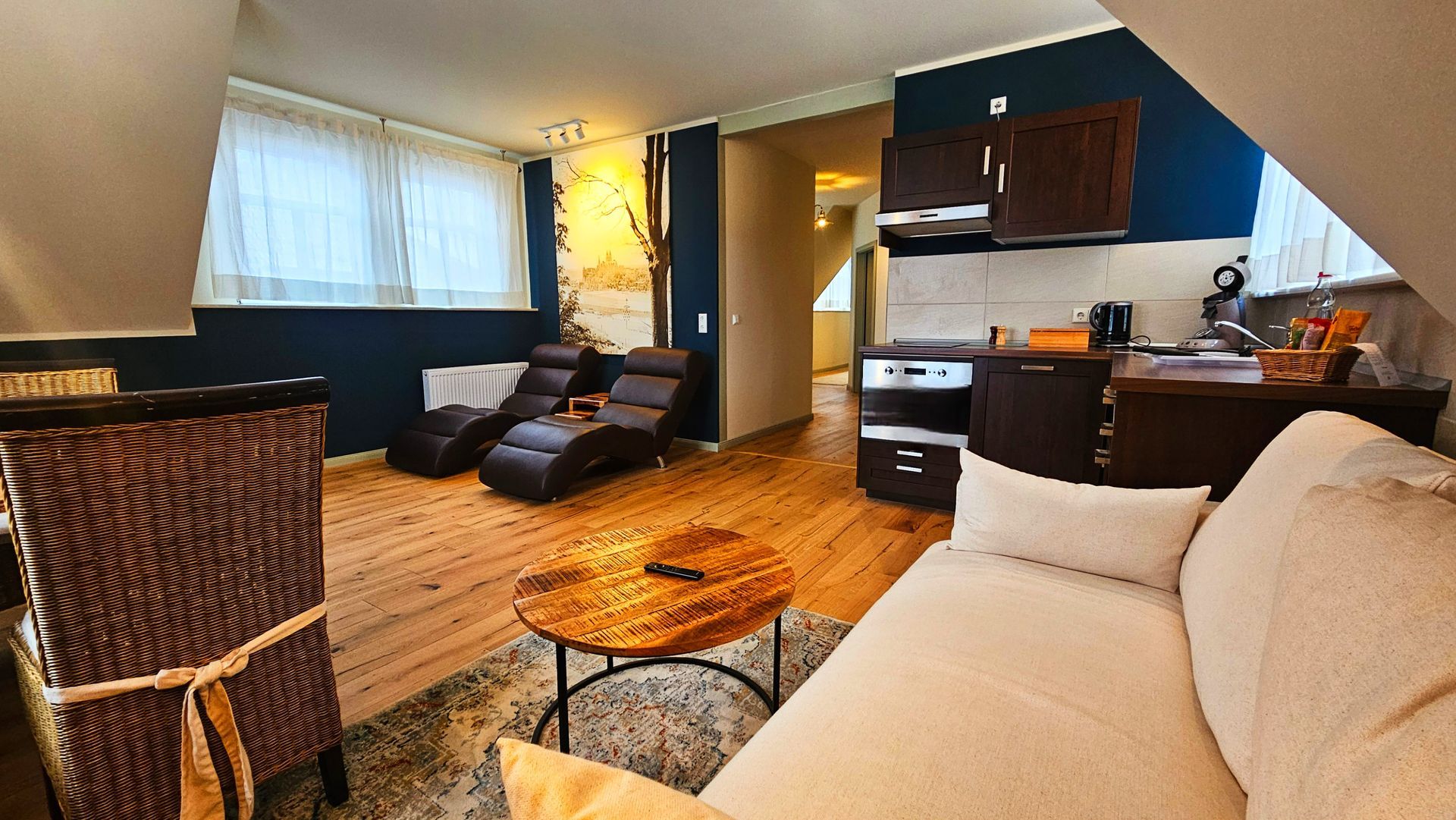 Gemütliches Dachzimmer mit Loungesesseln, kleiner Küche, Sofa und Holztisch auf einem Teppich.