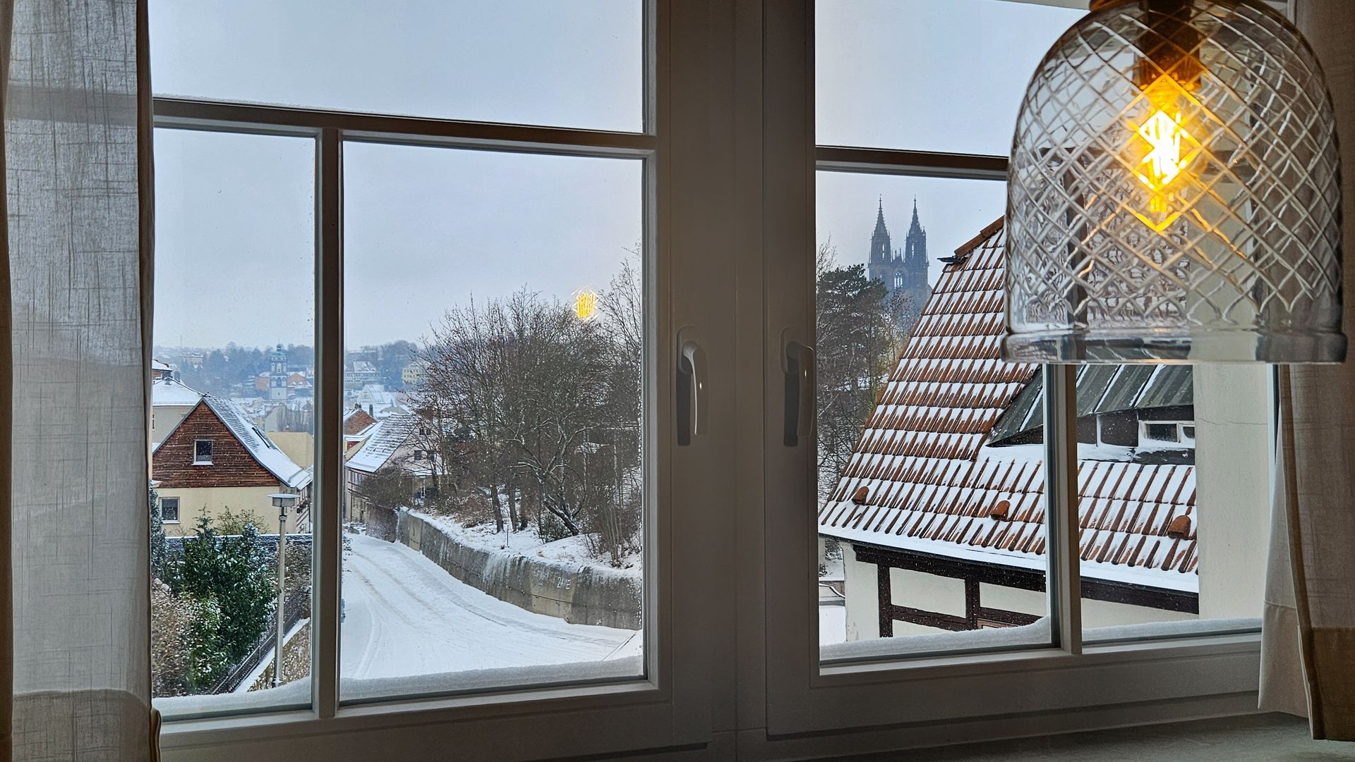 Blick durch ein Fenster auf verschneite Dächer und einen Kirchturm; drinnen hängt eine dekorative Lampe.