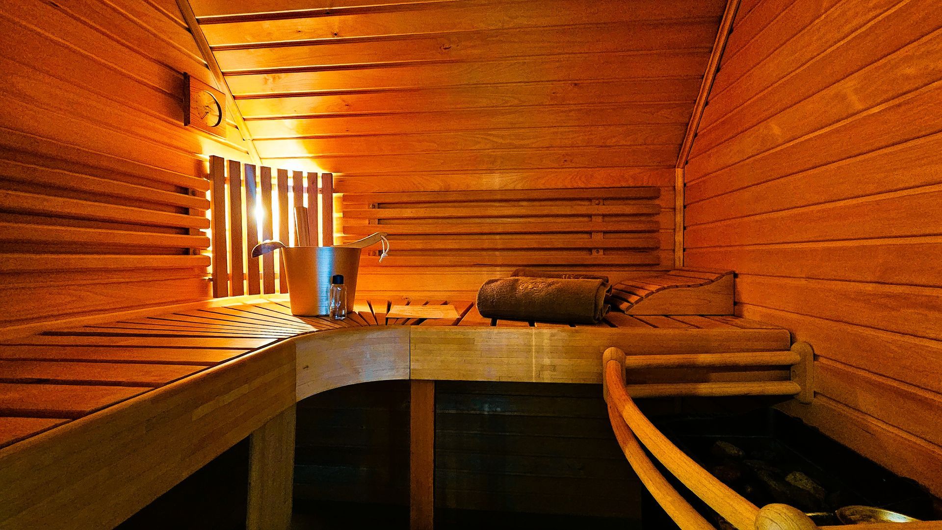 Saunainterieur aus Holz mit Bänken, Metalleimer, Heizung und warmer Beleuchtung.