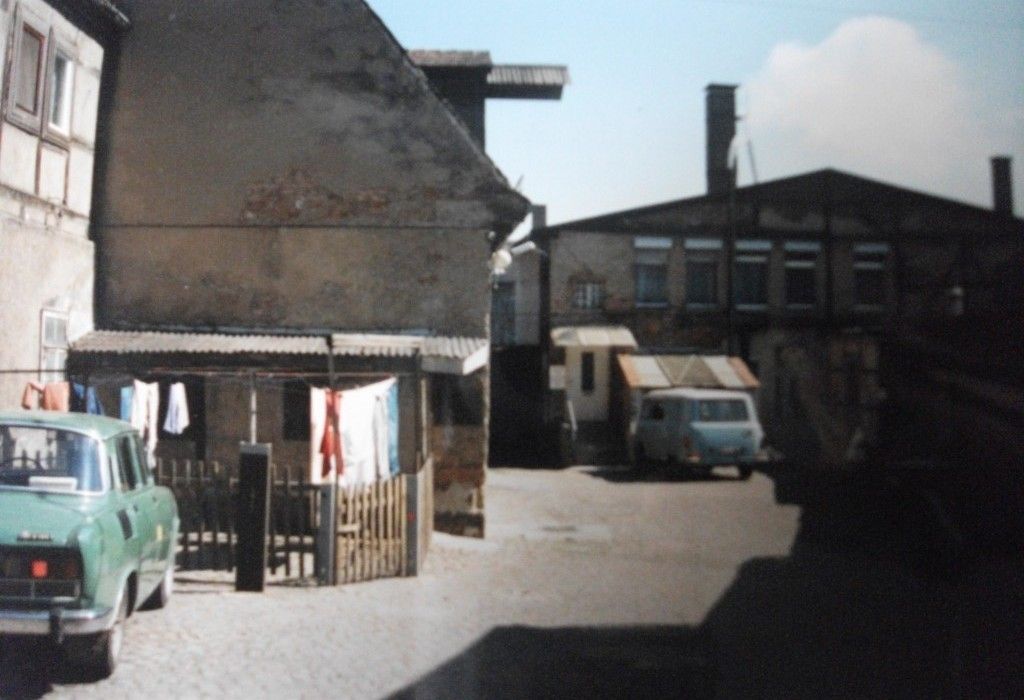 Alte Gebäude und ein Auto mit hängender Wäsche in einer engen Gasse. Ein Lieferwagen steht neben einem größeren Gebäude.