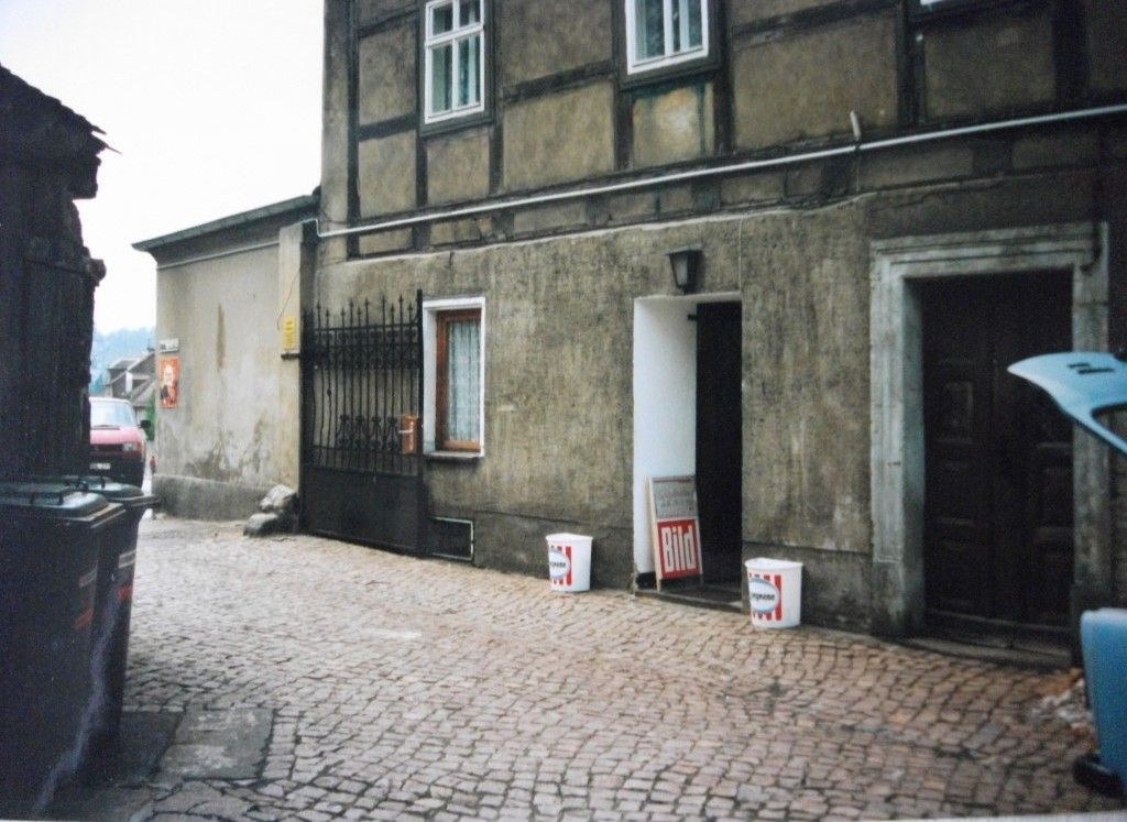 Kopfsteinpflasterstraße mit alten Gebäuden; vor einem Gebäude befindet sich ein dunkler Eingang und drei weiße Eimer.