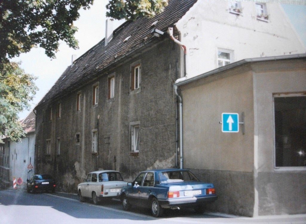 Straßenszene mit geparkten Autos neben einem langen, mehrstöckigen Gebäude und einem kleineren angrenzenden Bauwerk.