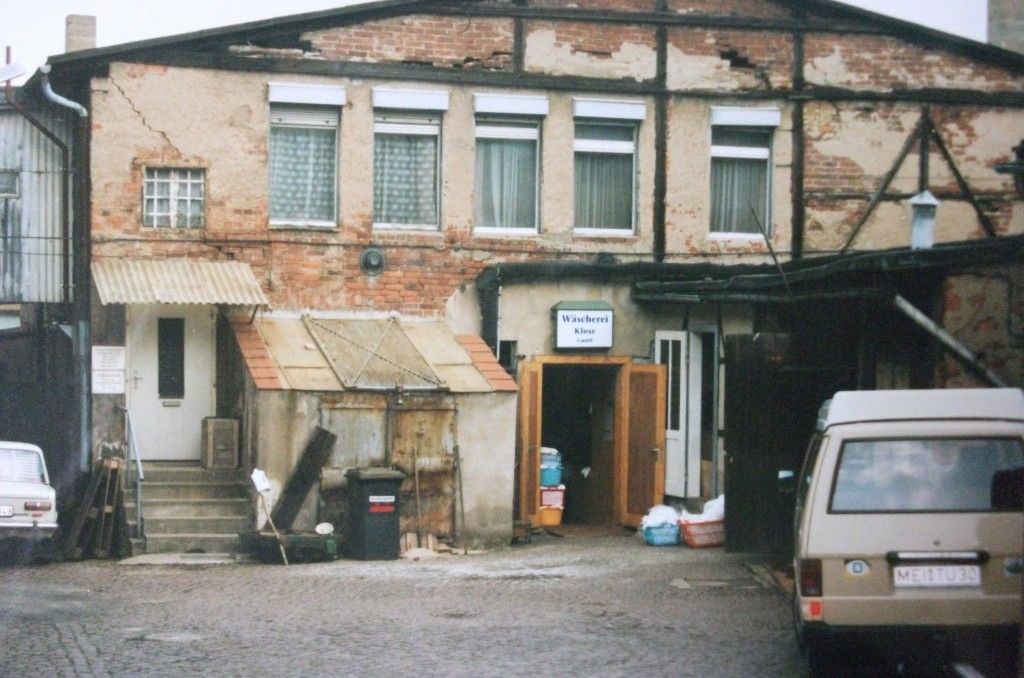 Ein heruntergekommenes zweistöckiges Gebäude mit vernagelten Fenstern und einem angebauten Schuppen. Ein geparkter Lieferwagen und ein Auto sind zu sehen.
