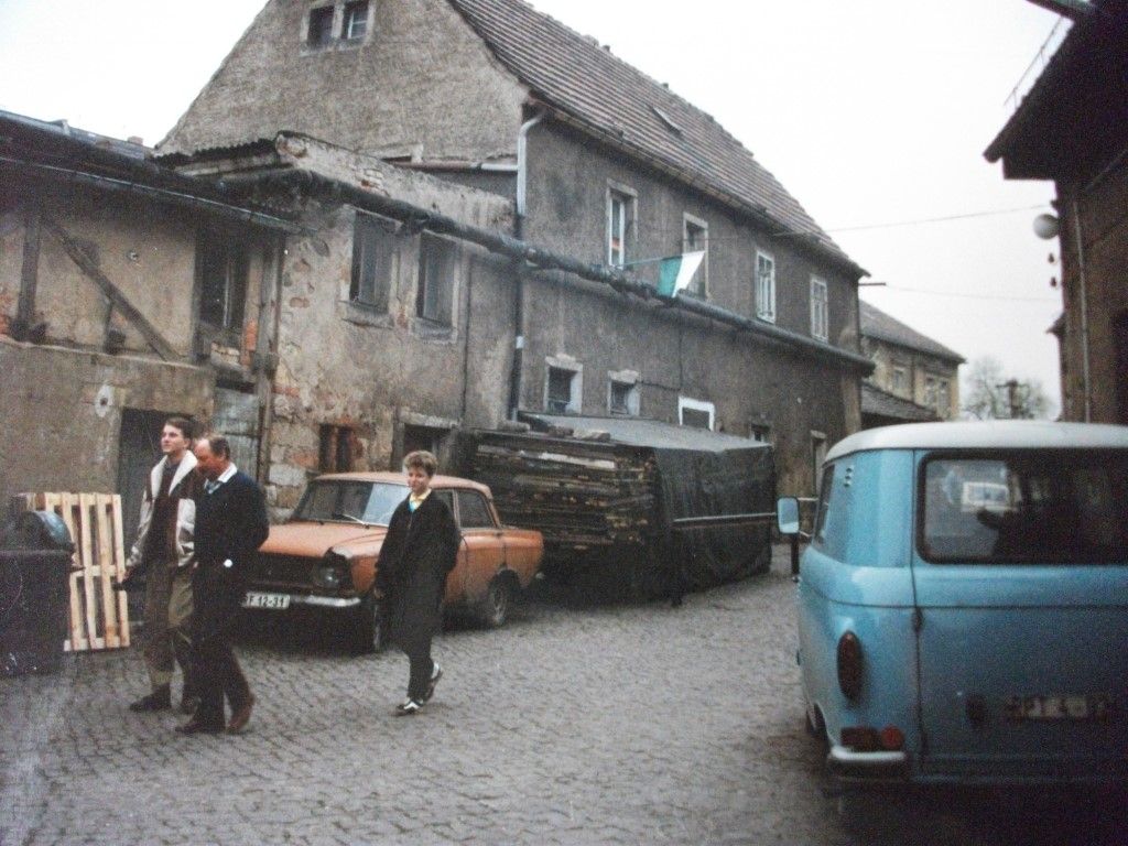 Kopfsteinpflasterstraße mit Menschen und geparkten Fahrzeugen. Verfallene Gebäude und ein dunkler Wohnwagen sind zu sehen.