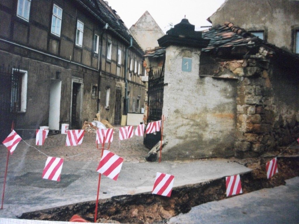 Straße mit rissigem Pflaster, rot-weiß gestreiften Fahnen und alten Gebäuden.