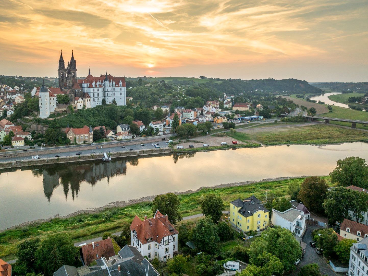 Stadtbild mit einer Burg auf einem Hügel, die sich im Fluss bei Sonnenuntergang spiegelt.