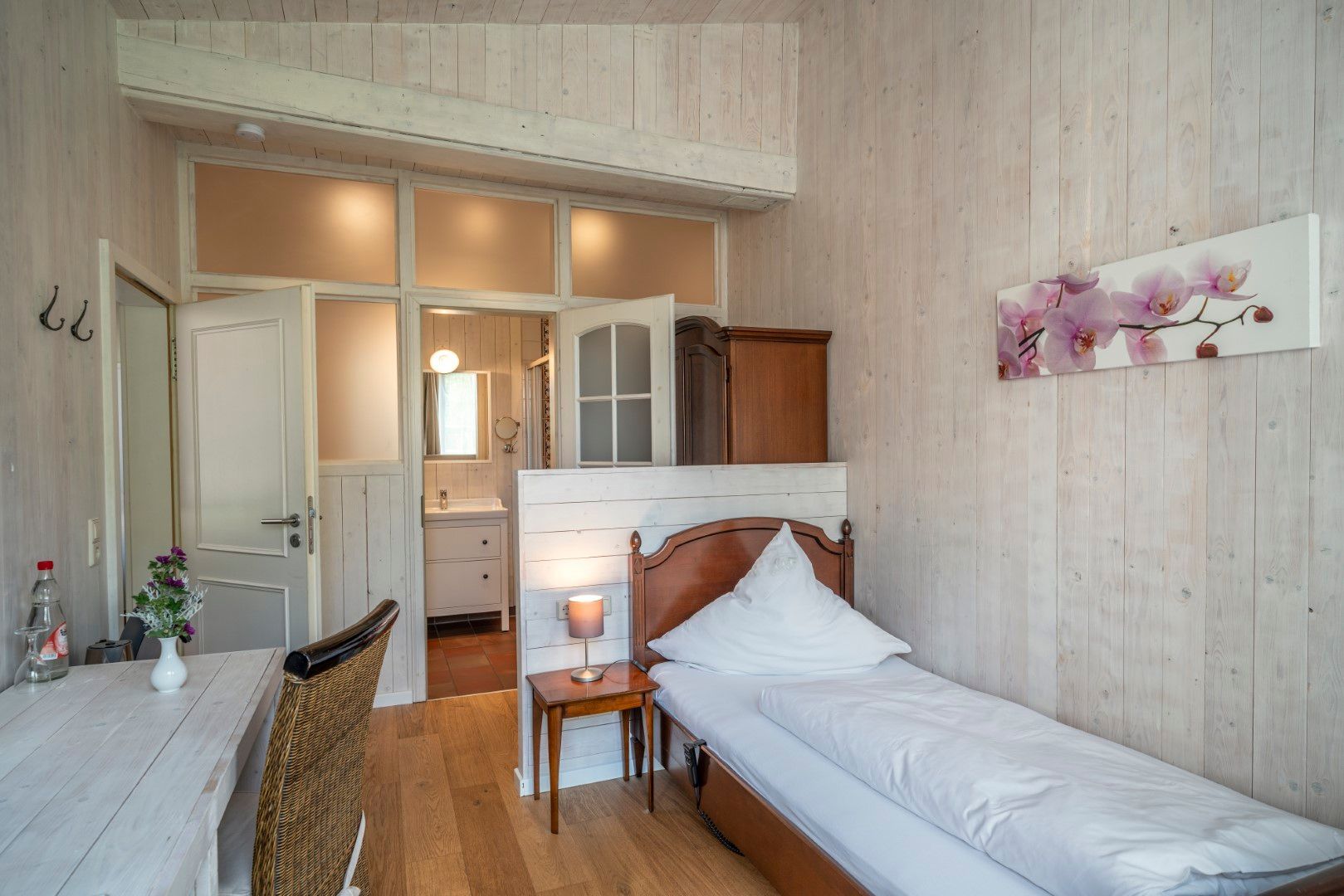 Schlafzimmer mit Einzelbett, Tisch und Zugang zum Badezimmer. Helle Holzvertäfelung und ein Blumenbild schmücken die Wände.