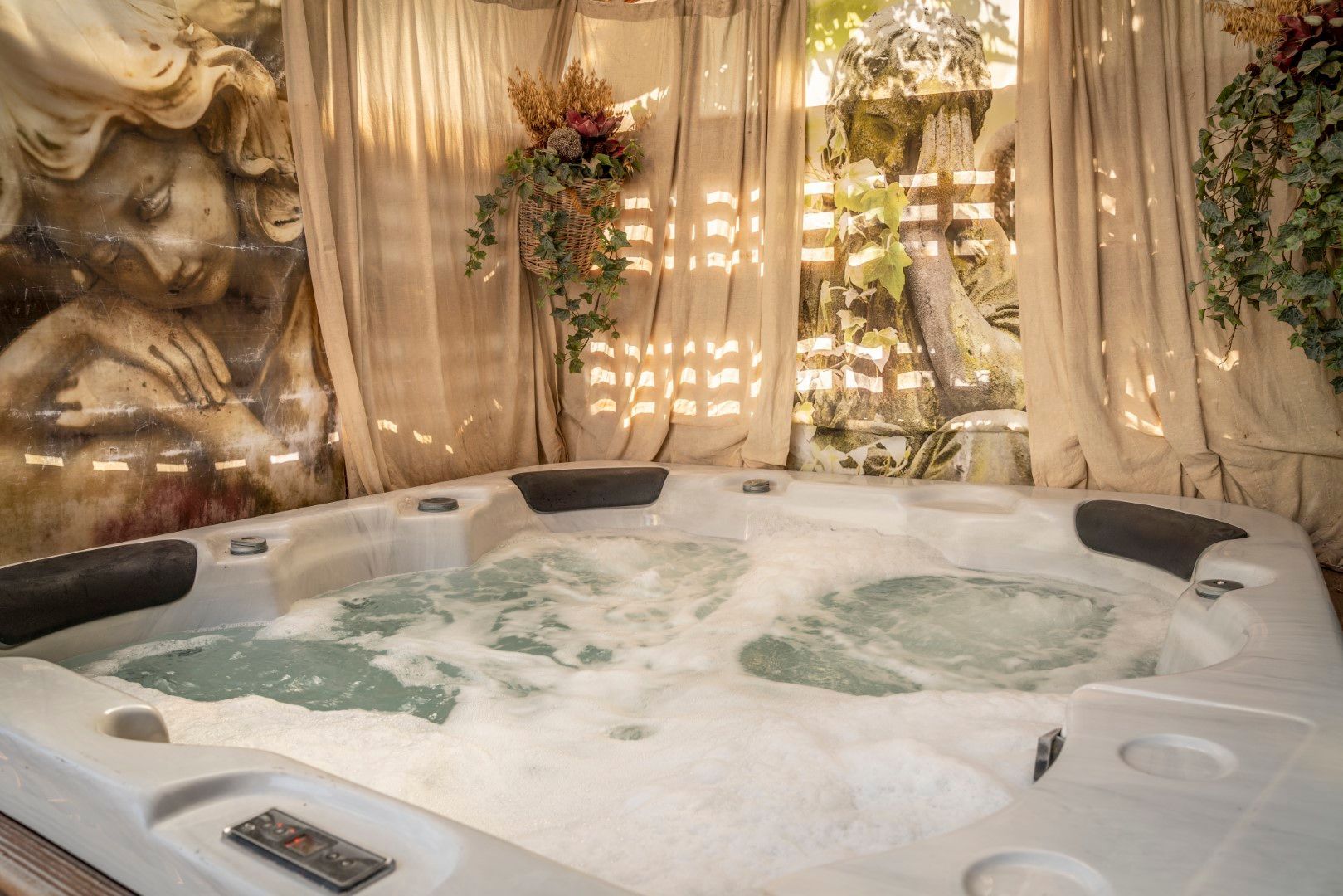 Whirlpool mit sprudelndem Wasser, eingebettet in einen Garten mit Statuen, drapierten Vorhängen und Hängepflanzen.