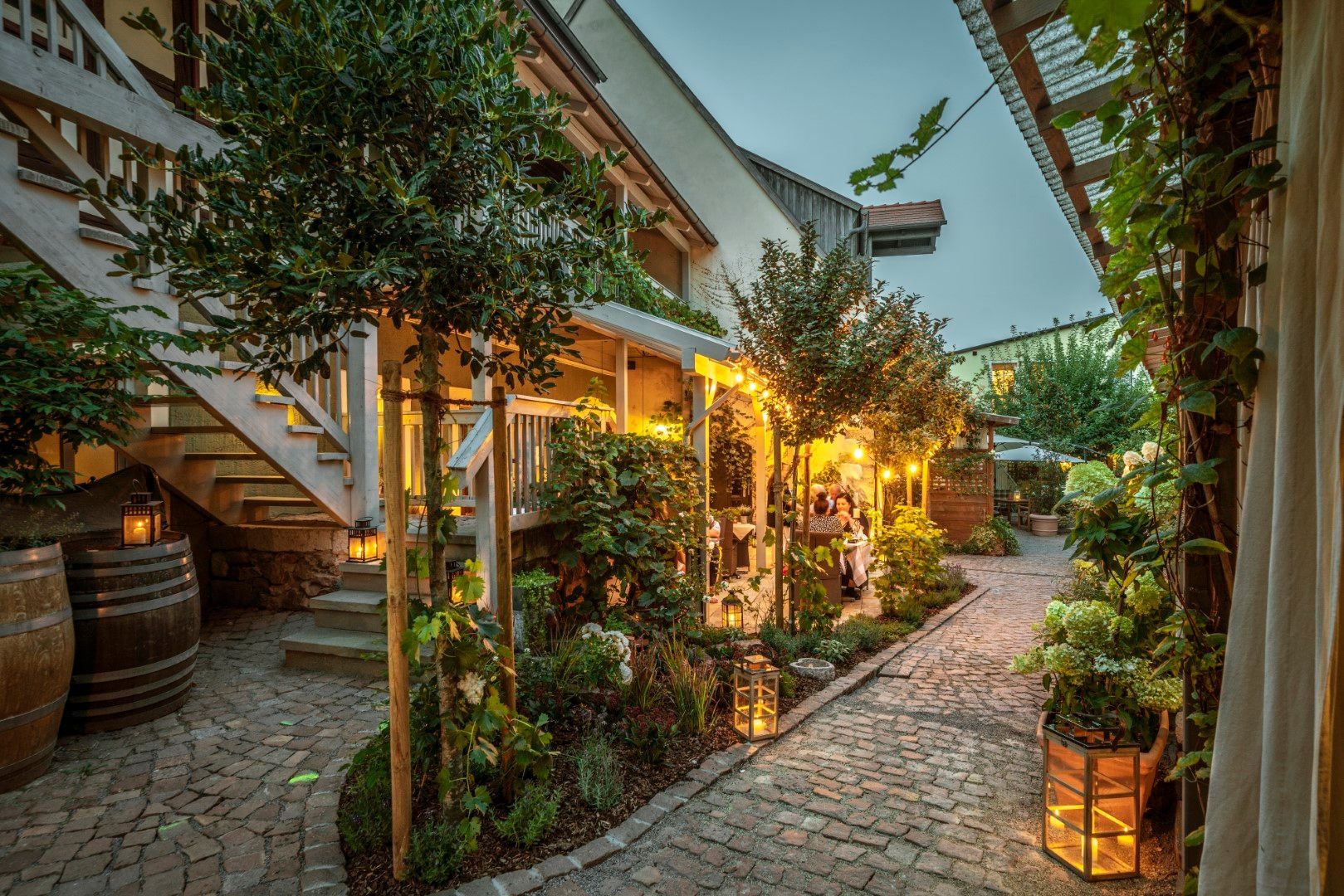 Innenhof mit Steinweg, beleuchtet von Laternen und Lichterketten, umgeben von Bäumen, Weinreben und Gebäuden.