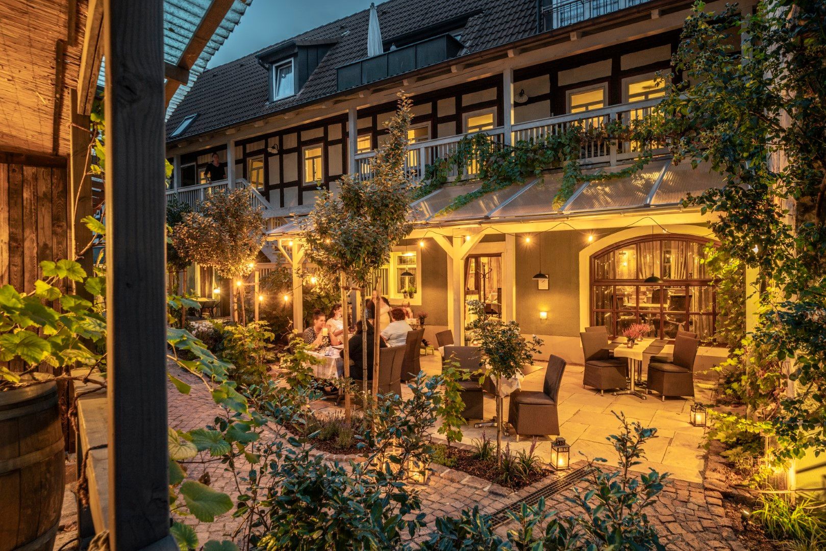 Das Restaurant im Innenhof bei Nacht, Gäste sitzen an Tischen. Lichterketten beleuchten das Fachwerkgebäude und das Grün.