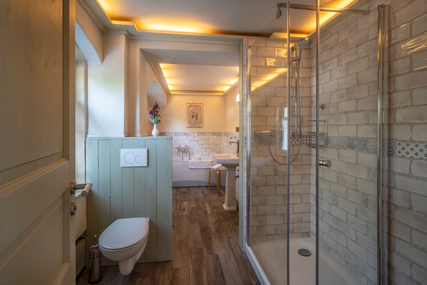 Badezimmer mit ebenerdiger Dusche, WC und Waschbecken. Holzboden, cremefarbene und graue Fliesen, warme Beleuchtung.